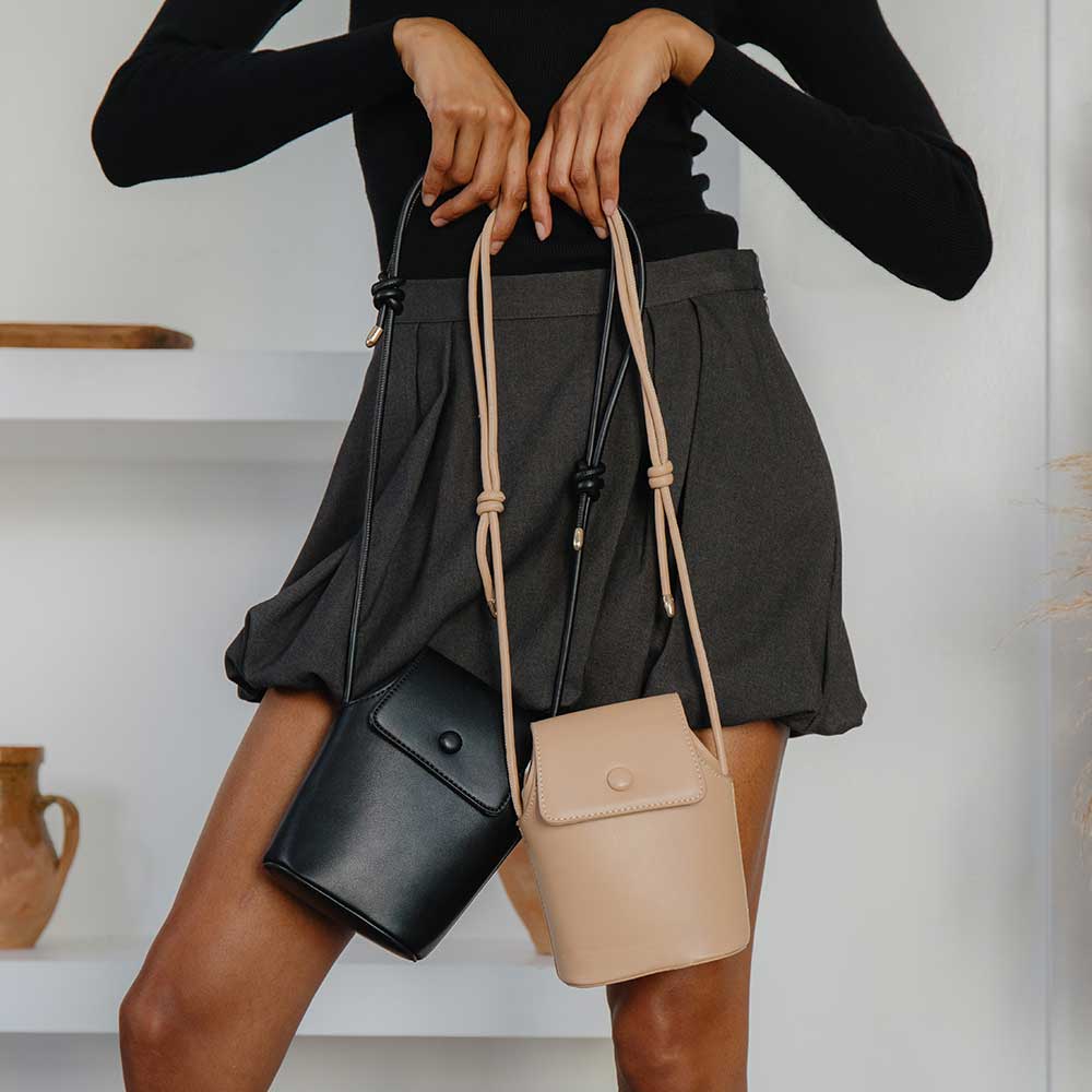 Person holding two handbags, one black and one beige, against a neutral background. Abigail Rust Red Crossbody Bag Melie Bianco