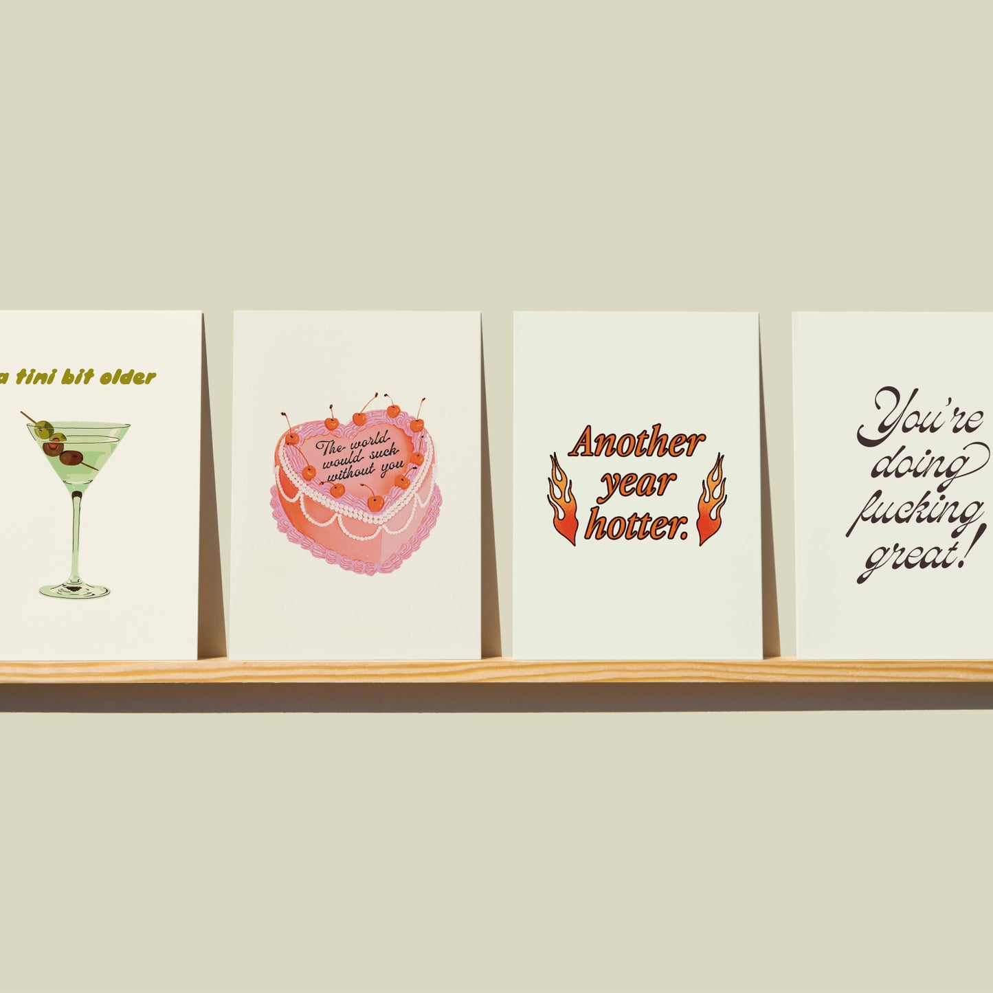 Four greeting cards on a wooden shelf against a beige wall.