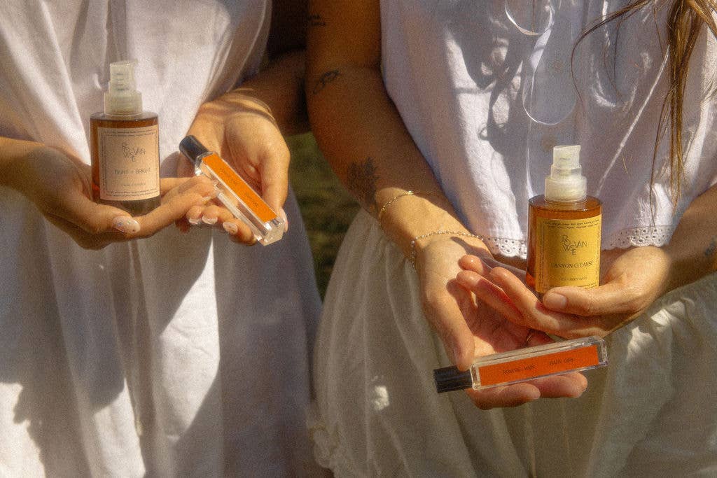Two people in white dresses outdoors holding small cosmetic bottles and tubes with orange liquid, under bright natural sunlight.