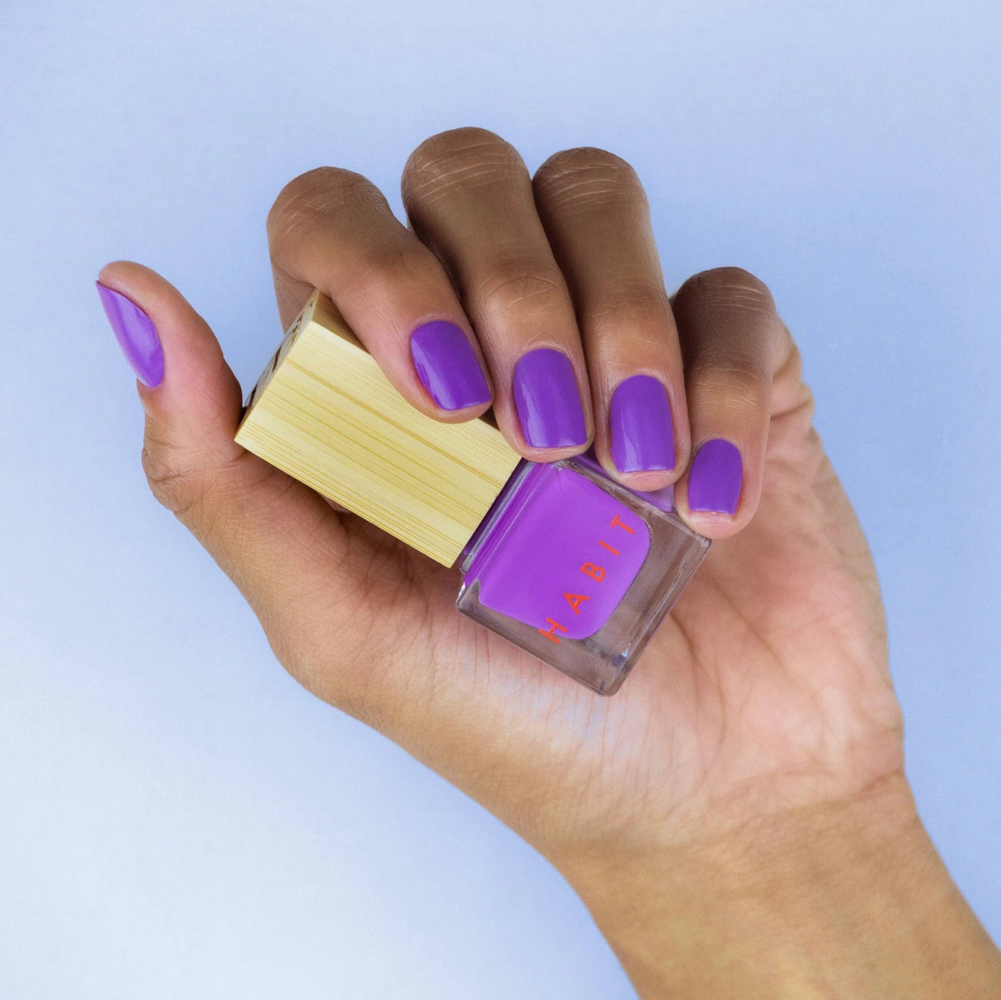 Hand holding a bottle of purple nail polish with matching nails against a light blue background, Belladonna Purple Toxin Free Nail Polish