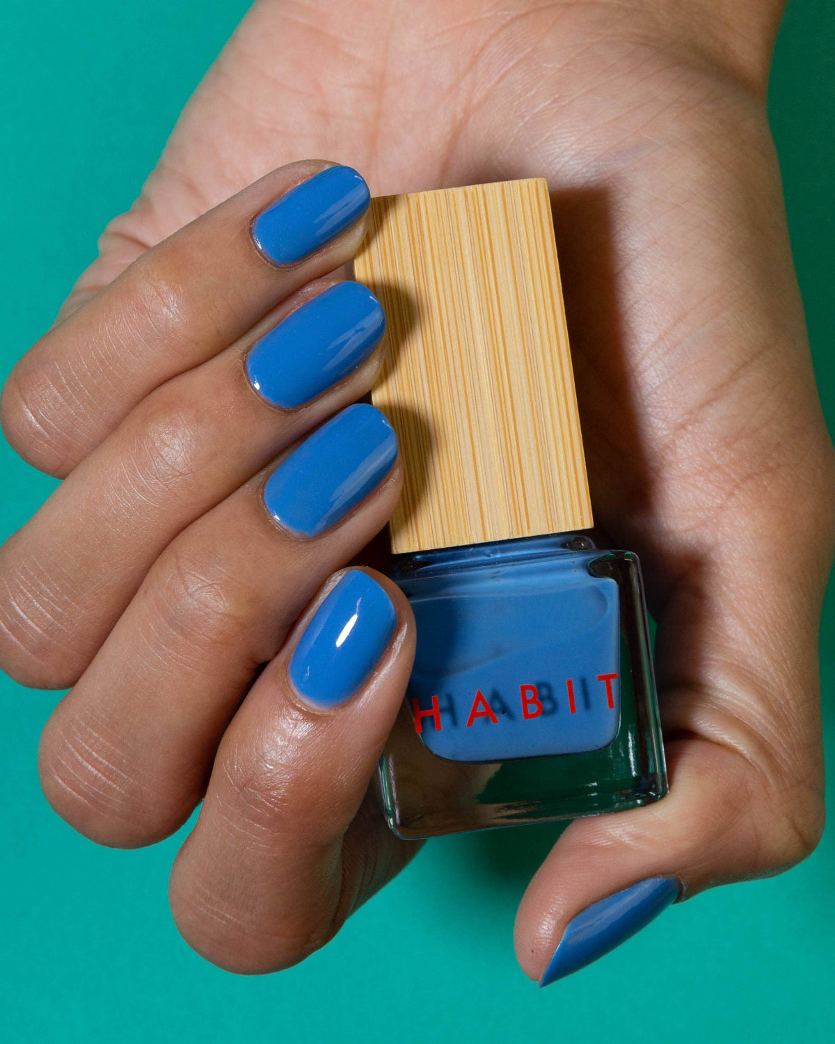 Hand holding a bottle of blue nail polish with 'Habit' branding against a teal background, Blue Jean Baby Blue Toxin Free Nail Polish