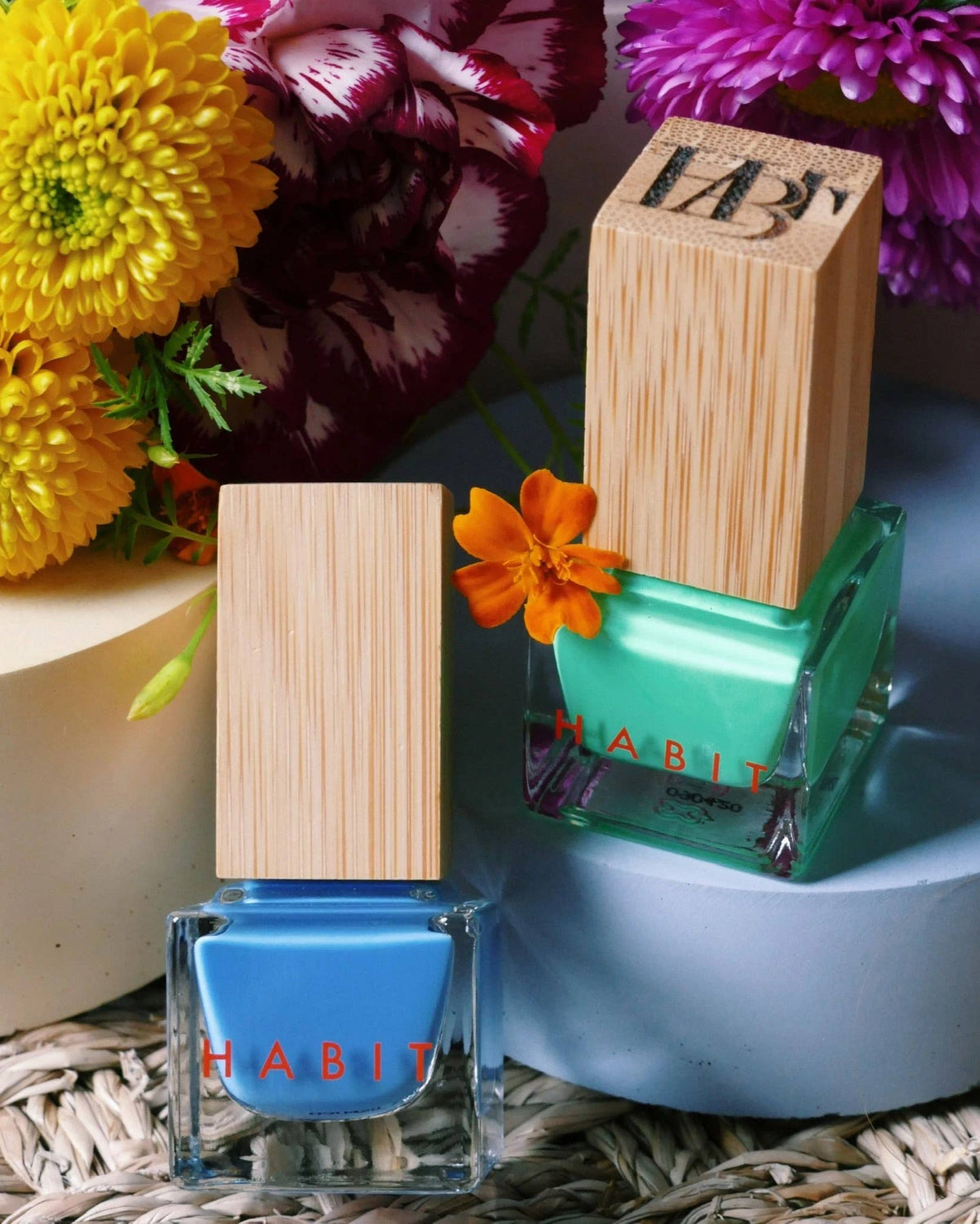 Two nail polish bottles with wooden caps on a decorative surface with flowers in the background. Blue Jean Baby Blue Toxin Free Nail Polish