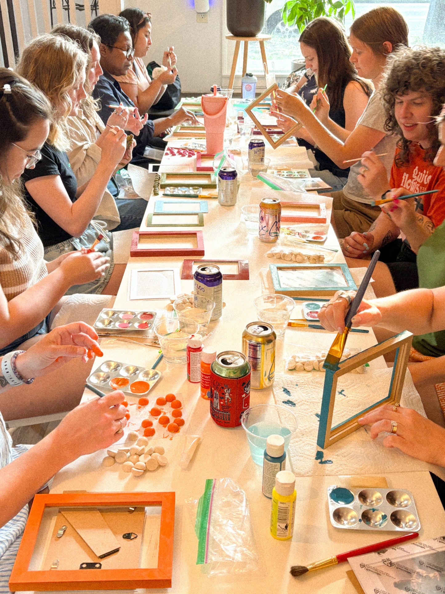 A group of people sitting around a long table, painting and assembling framed art pieces in a bright workshop or studio setting.