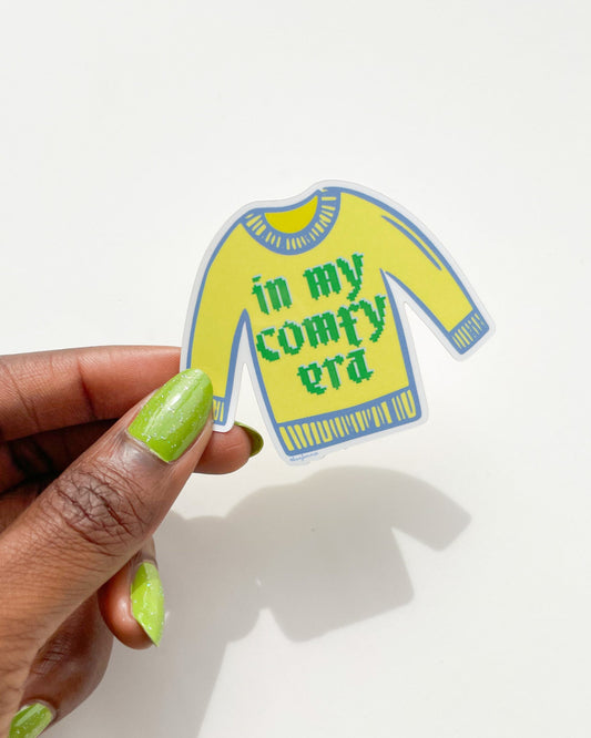 Sticker of a yellow sweater with text held by a hand on a white background, Comfy Era Sweater Sticker