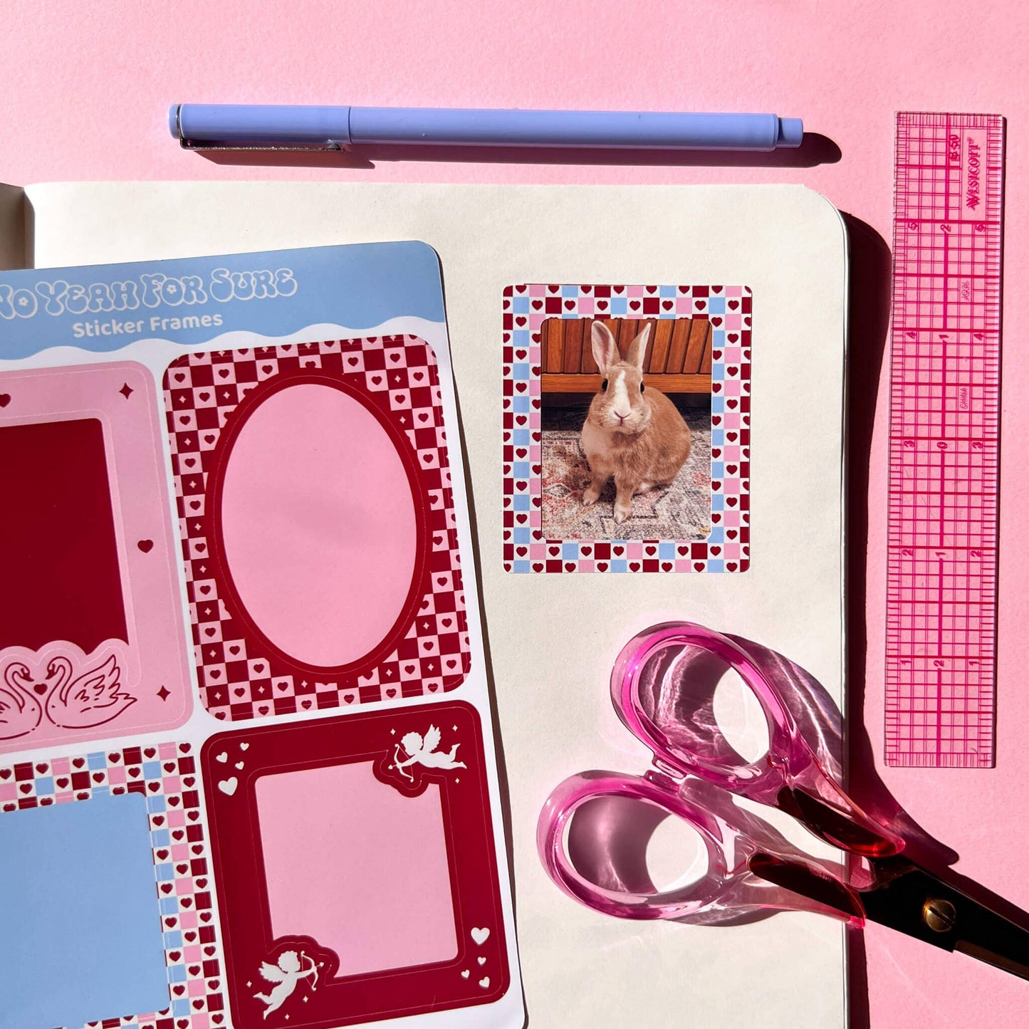 A styled flat-lay of sticker frames and crafting tools on a pink background. Visible items include a sheet of red, pink, and blue sticker frames, a sample sticker with a rabbit photo, a lavender pen, pink-handled scissors, and a pink ruler.