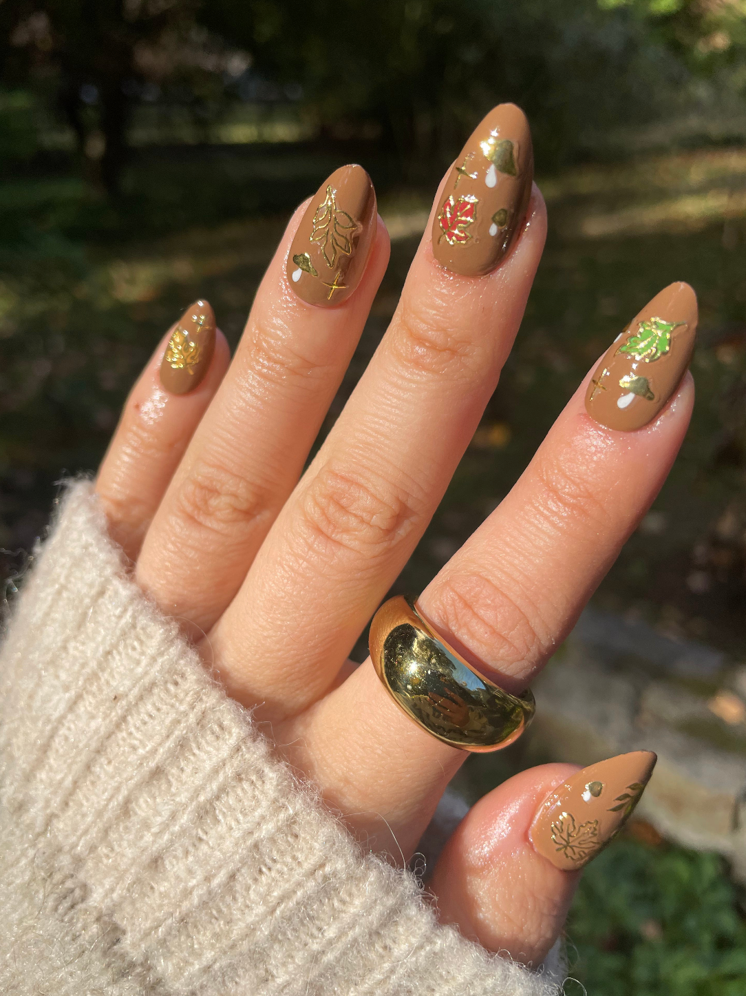 Hand with long, decorated nails and a gold ring, set against a blurred natural background. Cozy Fall Nail Art Stickers