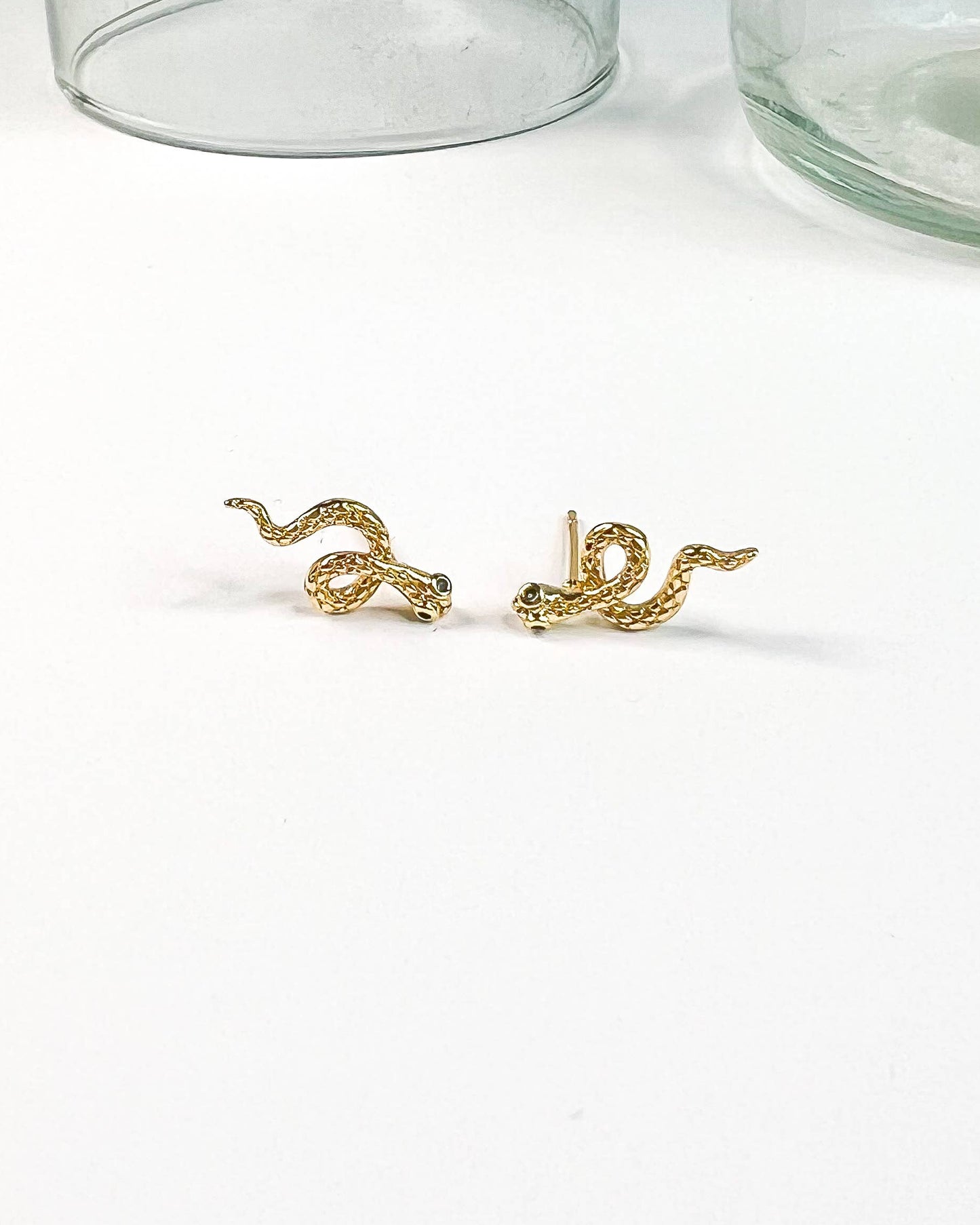 A pair of gold snake stud earrings displayed on a clean white surface, with two clear glass jars blurred in the background.
