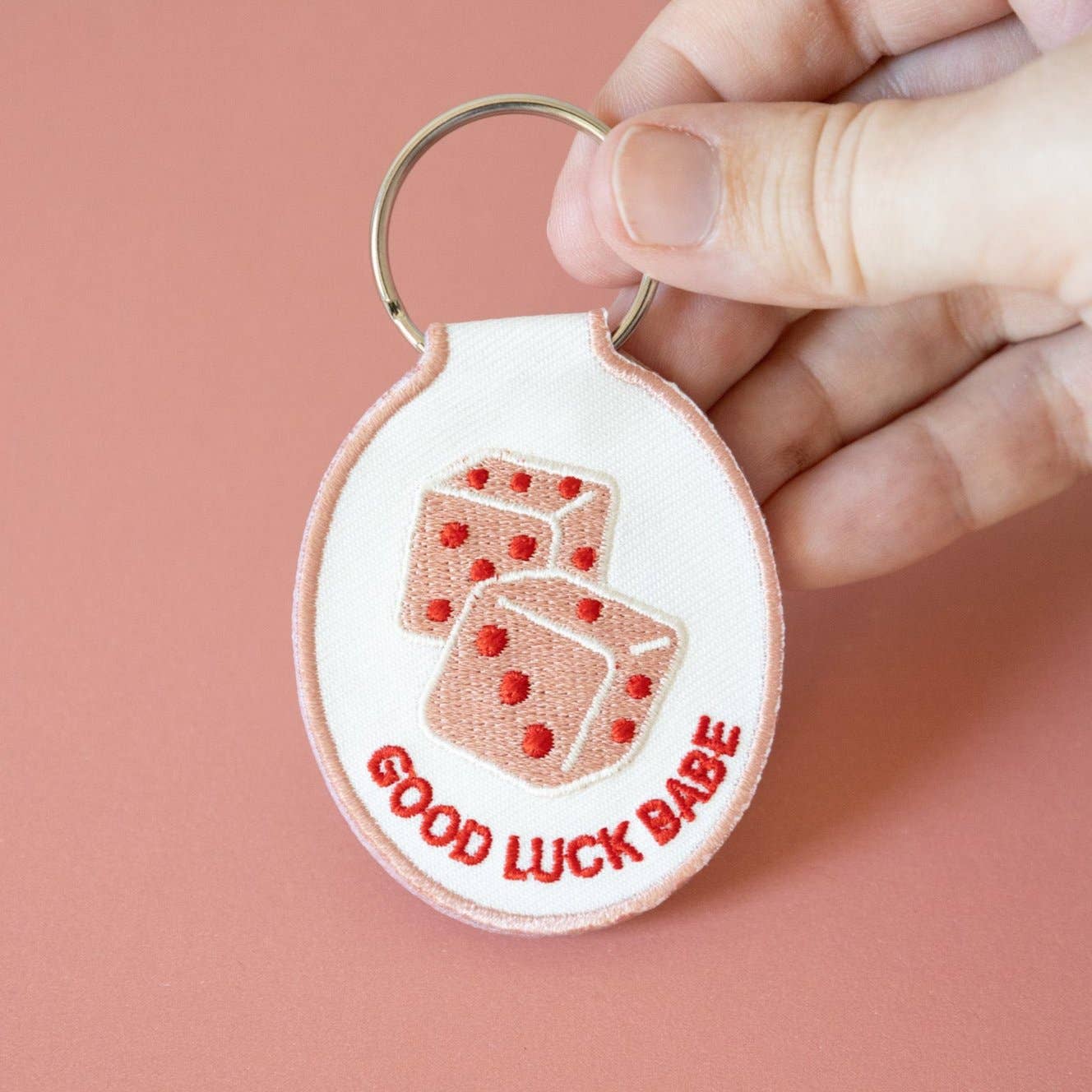 Keychain with dice design and 'Good Luck Babe' text held by a hand against a pink background