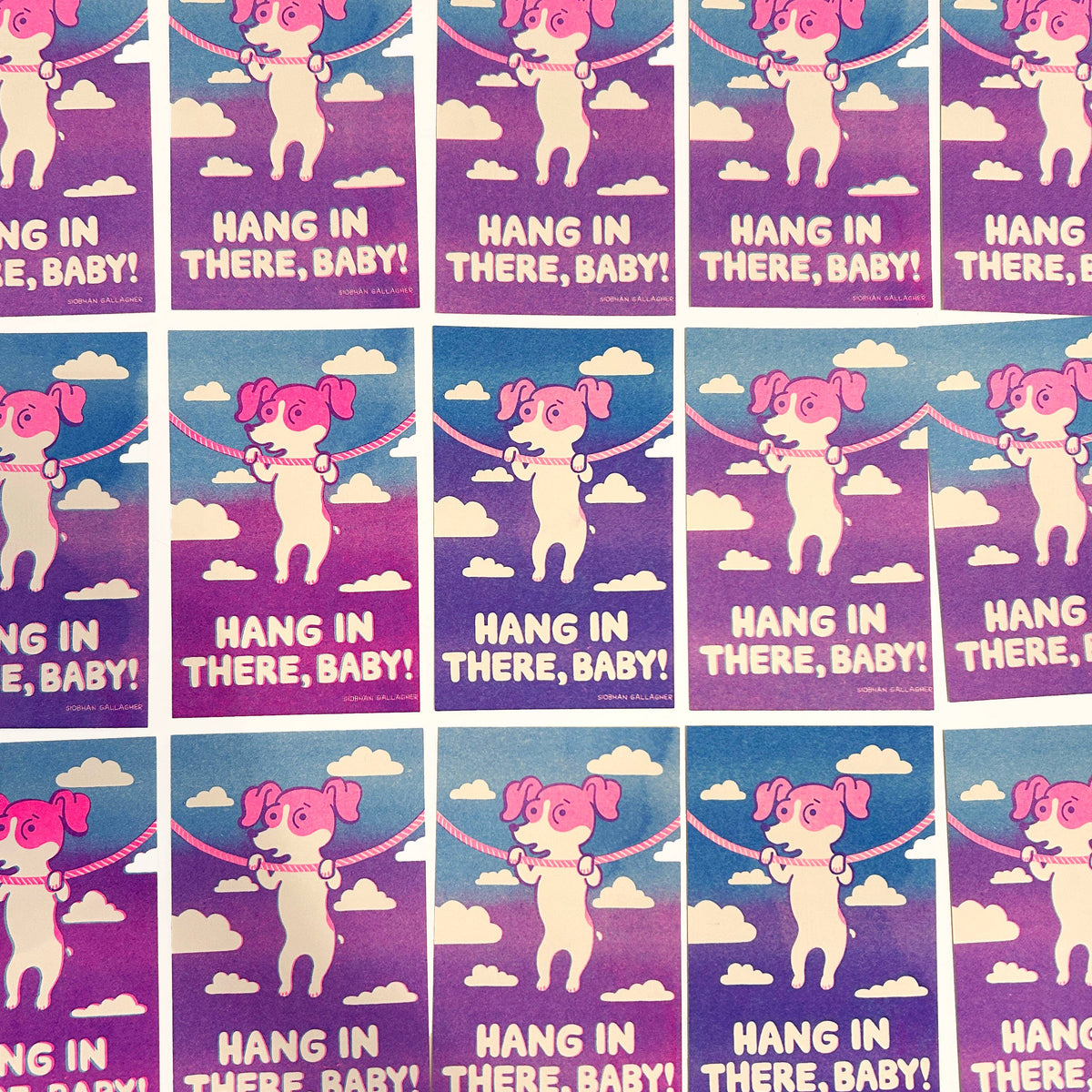 A repeating grid of art prints featuring a pink cartoon dog jumping on a rope against a purple-to-blue sky with clouds, and the text 'HANG IN THERE, BABY!' printed on each print.