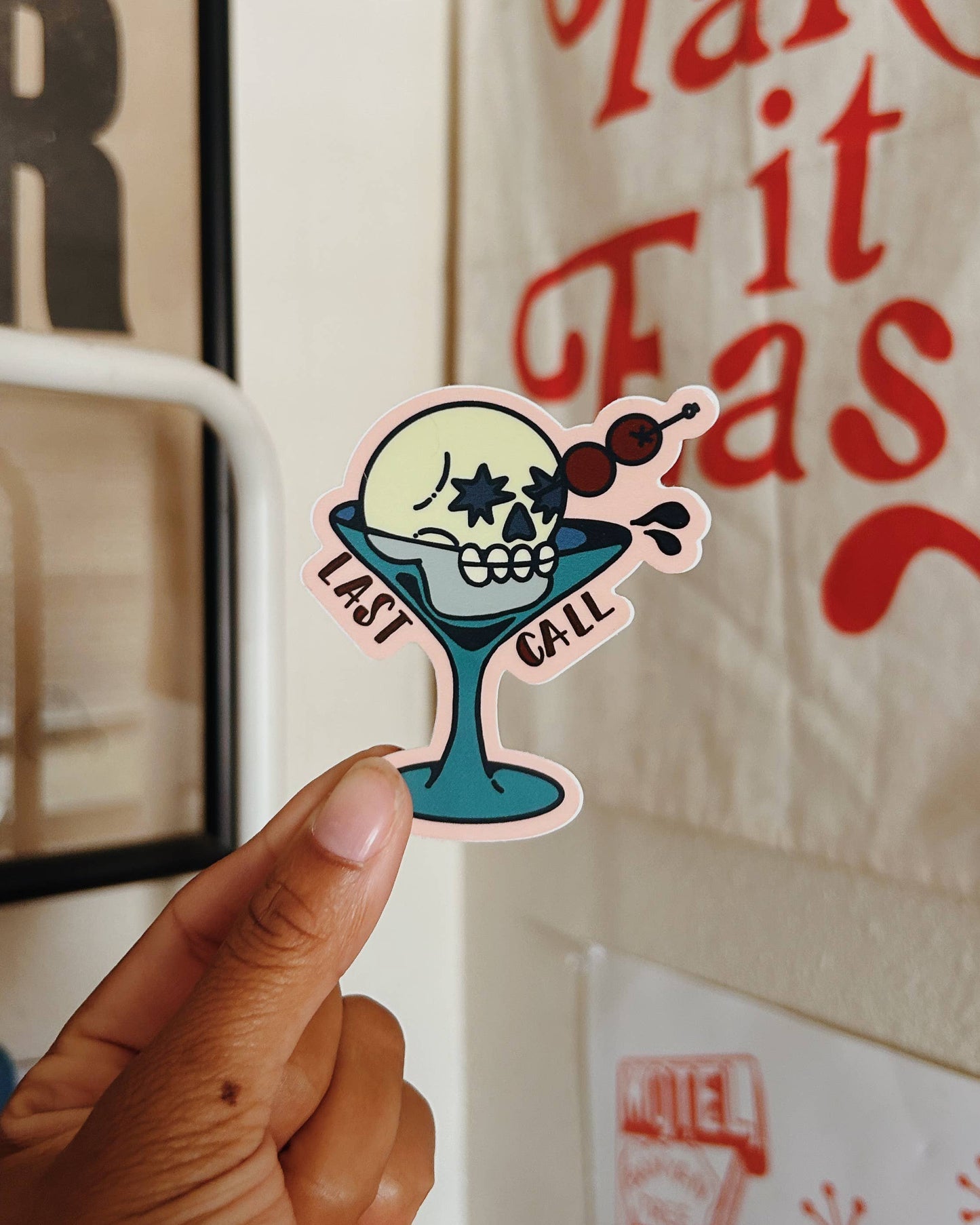 A hand holds a die-cut sticker featuring a cartoon skull in a teal cocktail glass with the words 'LAST CALL' , set against a background of a white wall with a canvas print and a lamp.