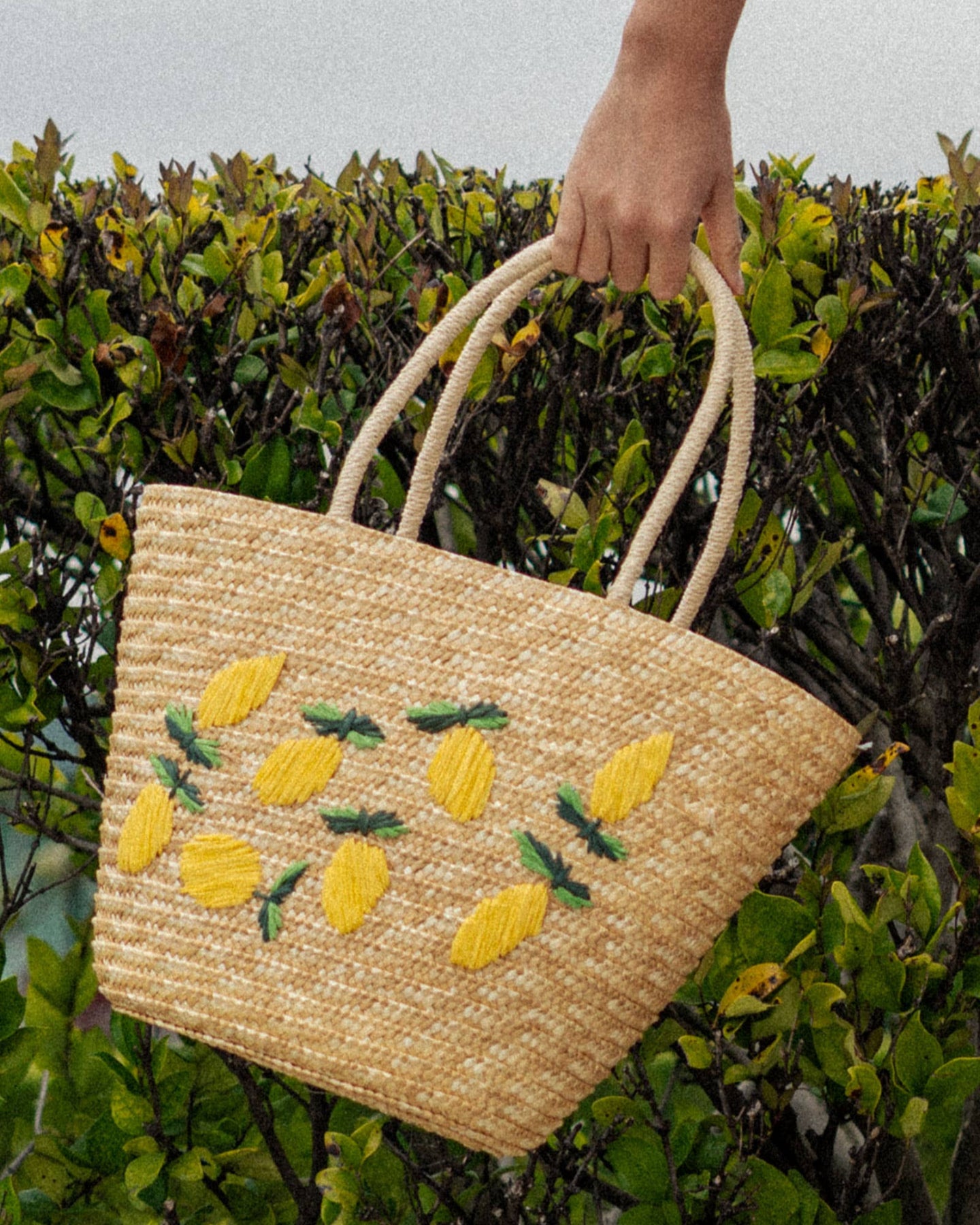 Woven handbag with lemon and pineapple patterns held by a person against a green leafy background