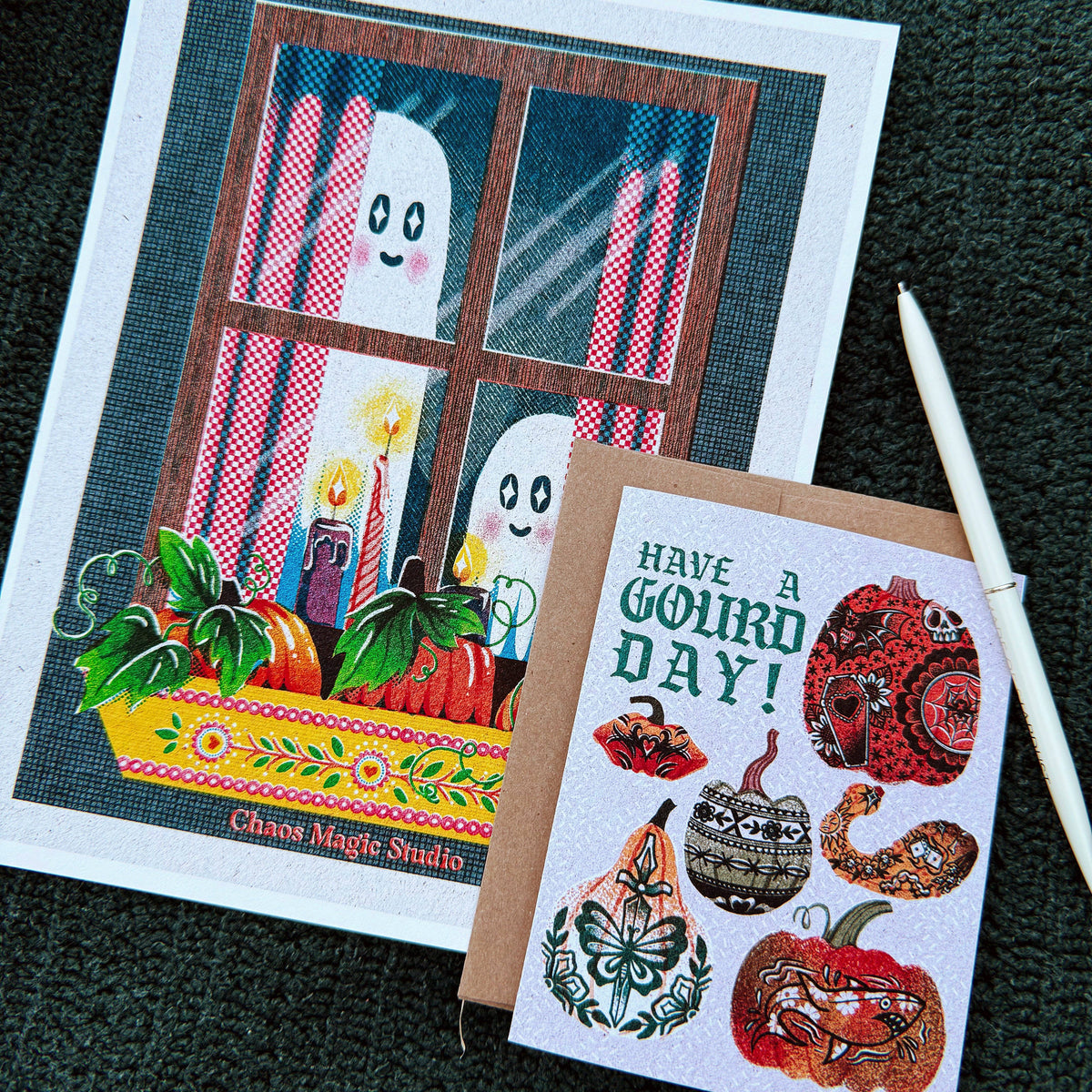Two illustrated greeting cards laid on a dark textured surface. The larger card shows a window scene with ghost figures, candles, pumpkins, and the text 'Chaos Magic Studio.