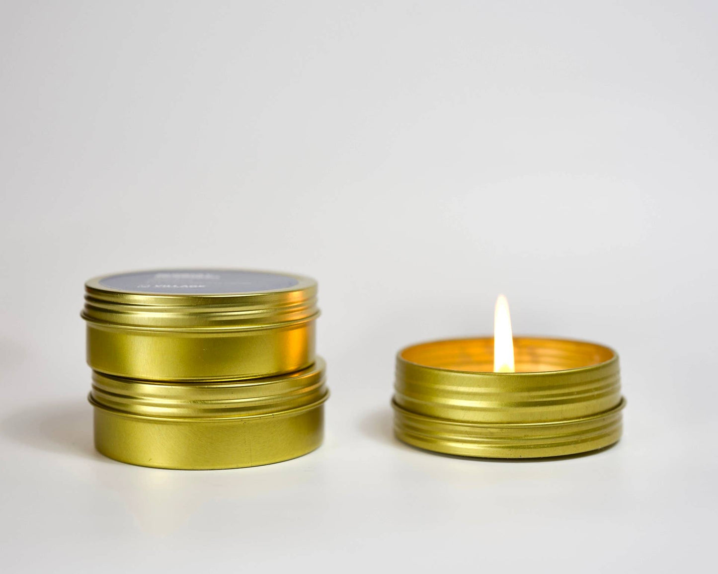 Two gold tins, one with a lit candle inside, on a light gray background