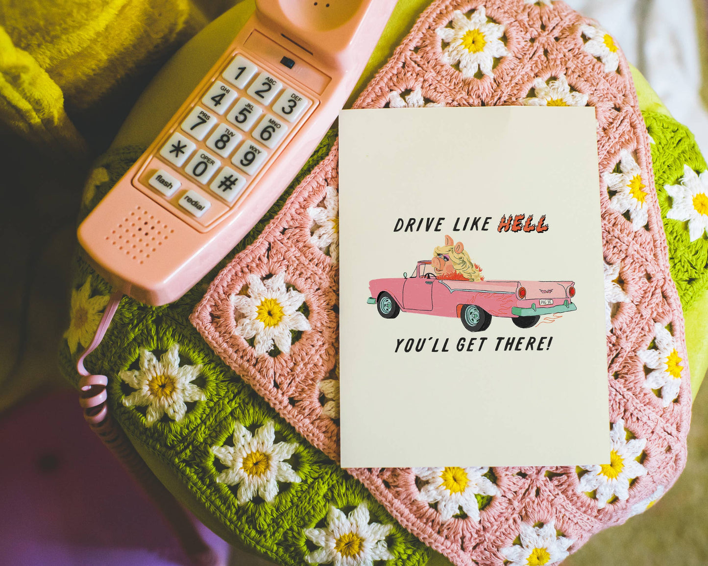 Art print with 'Drive Like Hell, You'll Get There!' text on a pink phone and crocheted surface with flowers.