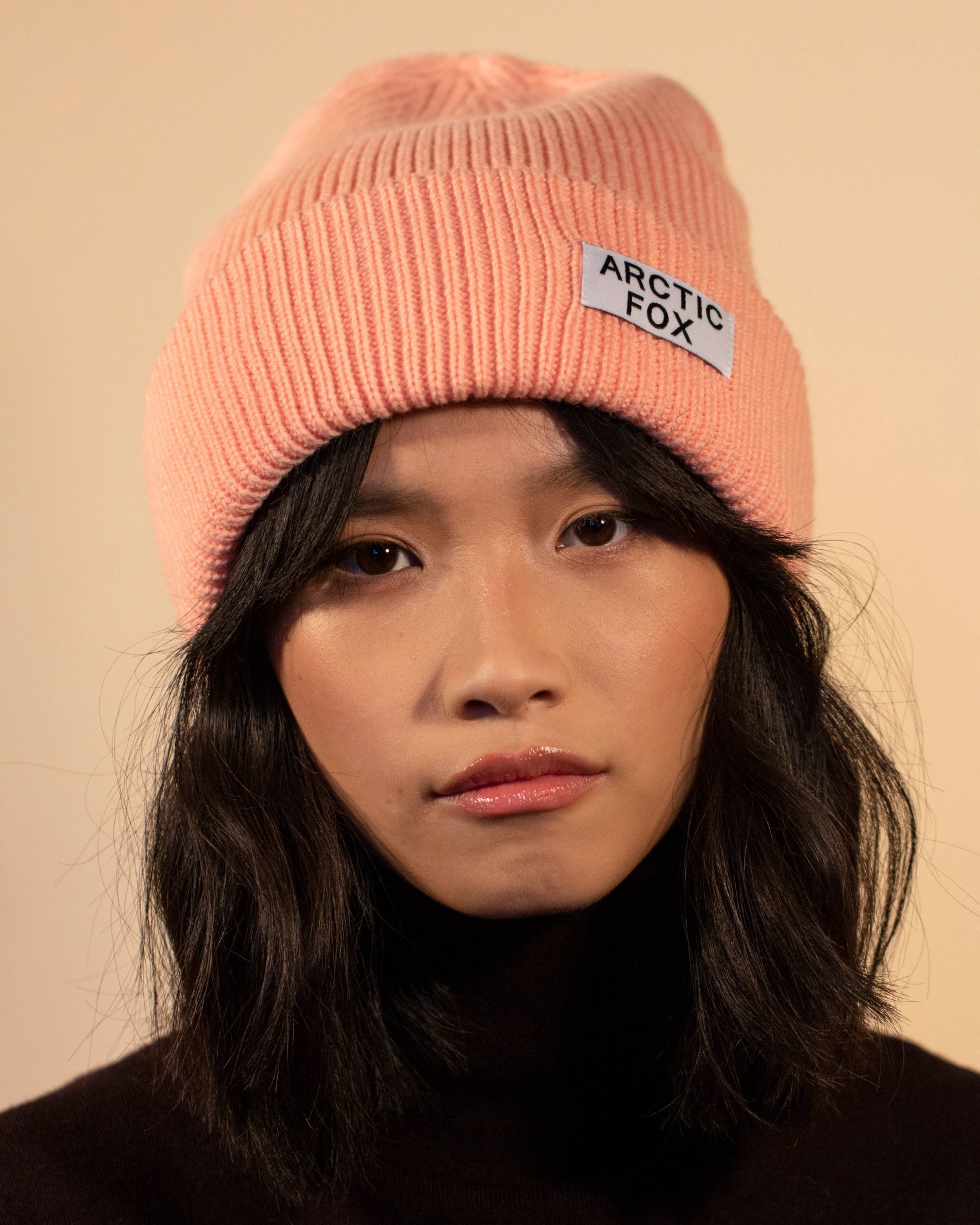 Person wearing a pink beanie with 'Arctic Fox' logo against a beige, Pink Recycled Bottle Beaniebackground