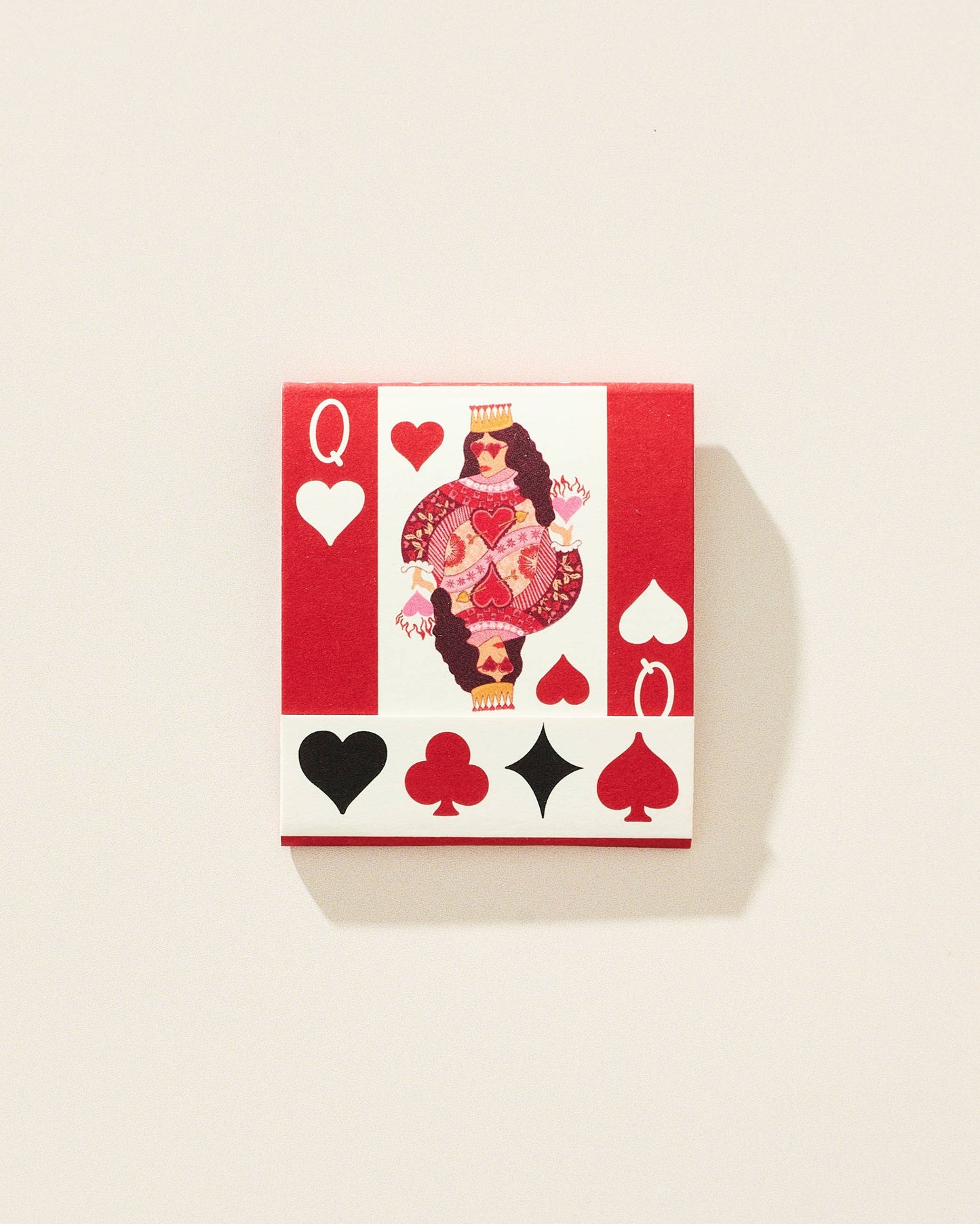 A square decorative matchbook featuring a stylized Queen of Hearts design with red and white background, black and red suit symbols, and a central illustrated figure, shown on a light beige wall with a soft shadow.