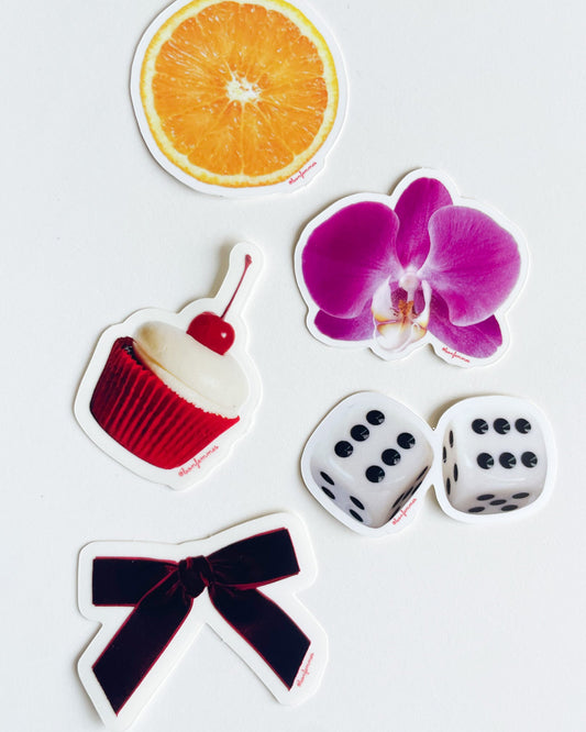 Set of five stickers featuring a cupcake, dice, bow, orange slice and flower on a white background.