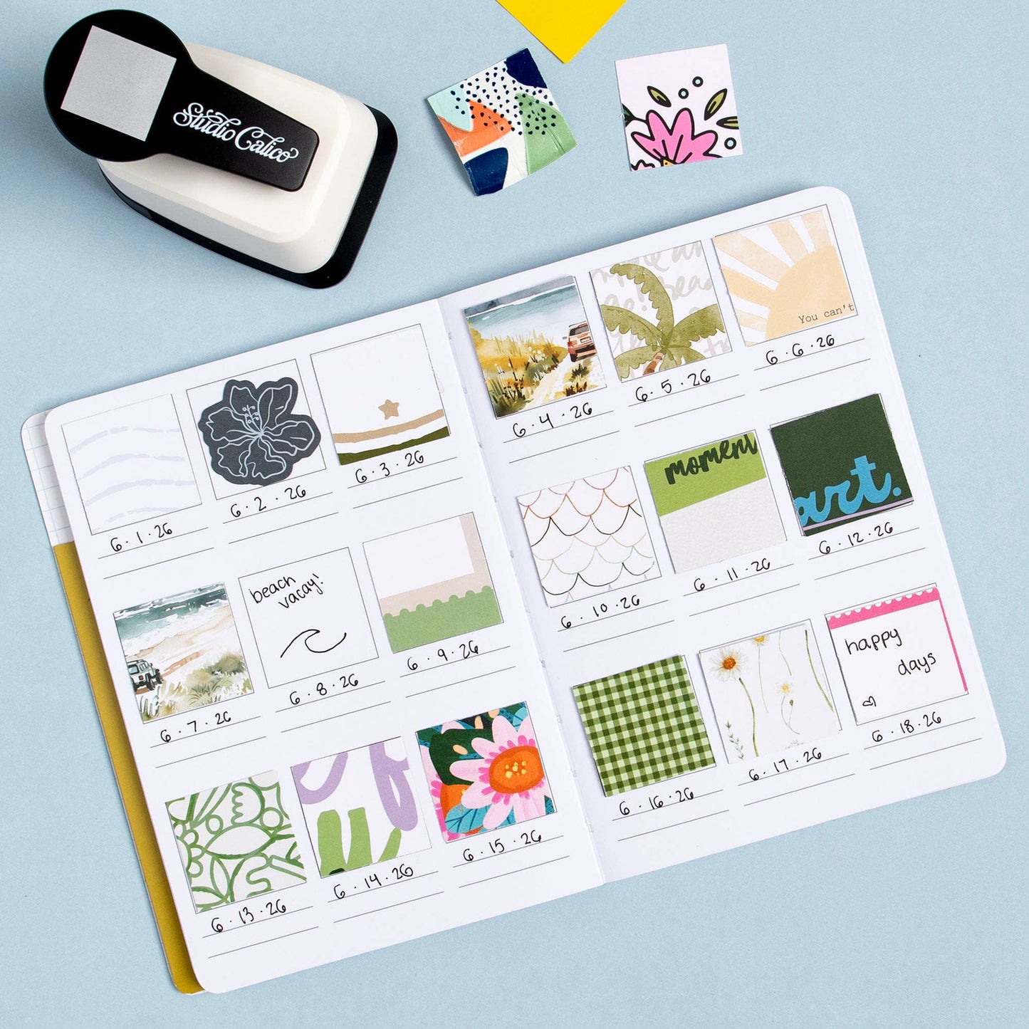 An open planner or notebook spread filled with small decorative stickers and notes, photographed on a light blue background. A black-and-white die-cut label maker and several sample sticker sheets are placed around it.