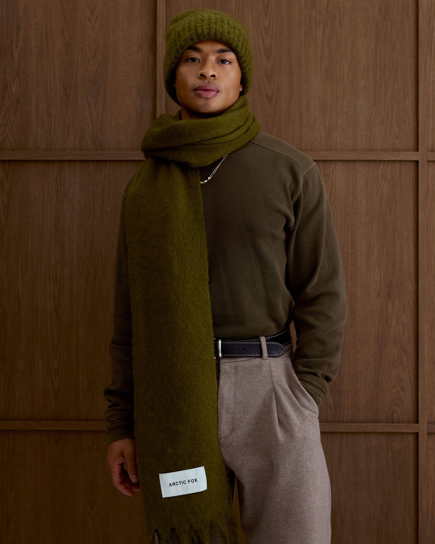 Person wearing a green scarf and hat against a wooden paneled wall, Sea Kelp Green Reykjavik Recyled Bottle Scarf