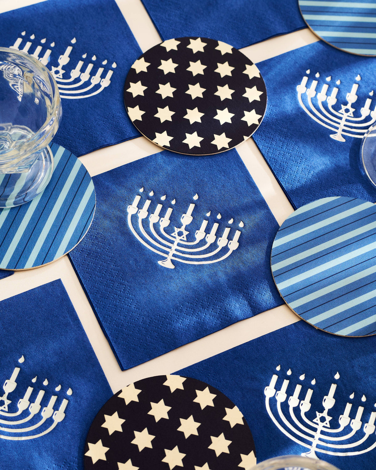 Blue and navy Hanukkah tableware set featuring square napkins with white menorah prints and round coasters in star and stripe patterns, styled with a clear glass on a cream lattice background.