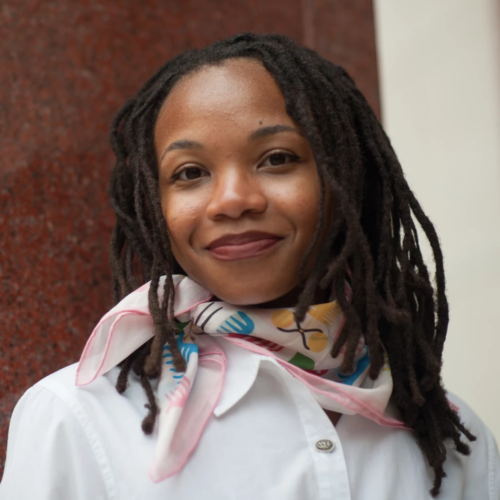 Woman with dreadlocks wearing a colorful scarf and white shirt against a red and beige wall