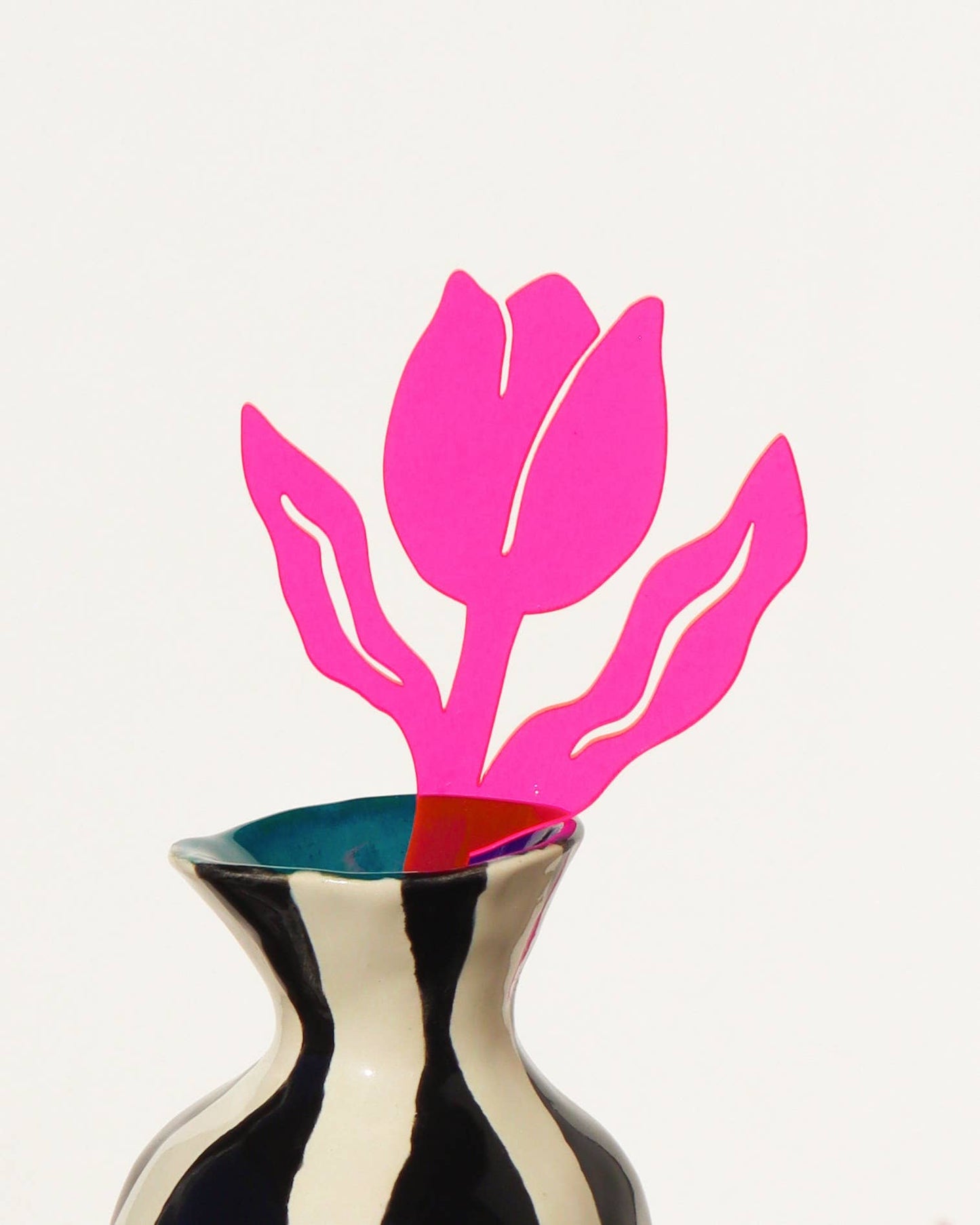 A bright pink tulip cutout decoration inserted into a black and white striped ceramic vase with a blue interior, set against a plain off-white background.