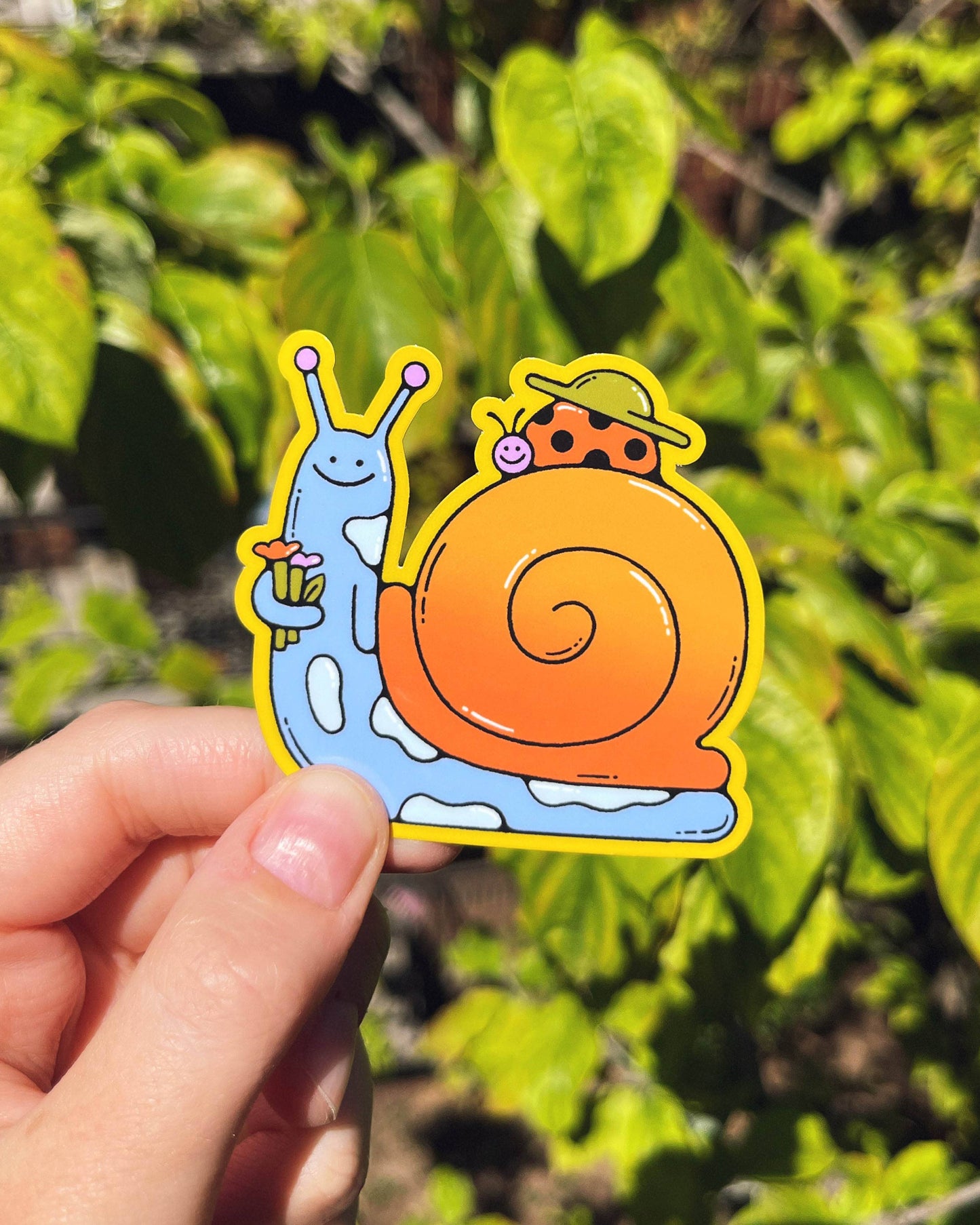 Hand holding a colorful sticker of two snails against a green leafy background