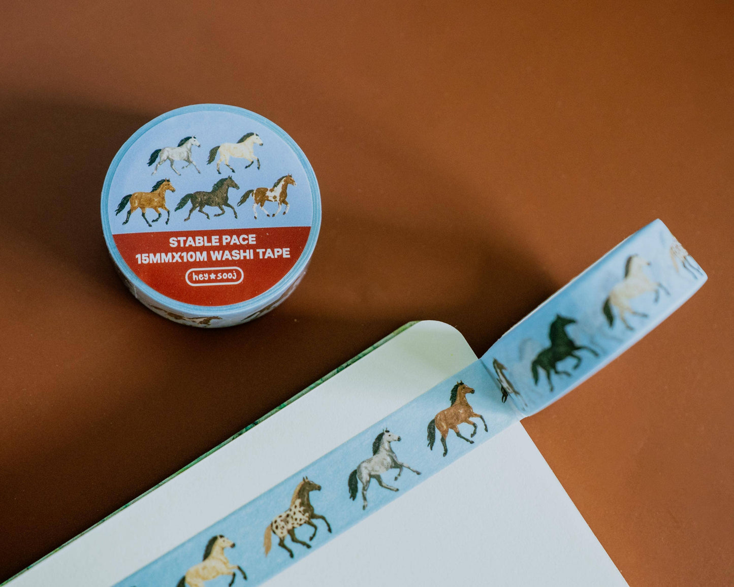A roll of washi tape with a light blue background and a pattern of running horses in black, brown, and white, shown next to an open notebook on a warm brown surface.