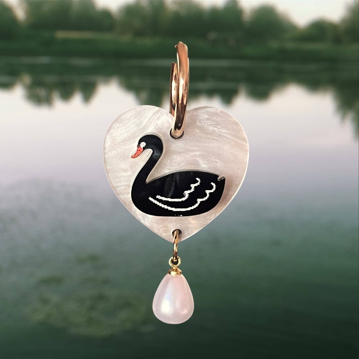 Heart-shaped pendant with swan design and pearl drop against a blurred natural background