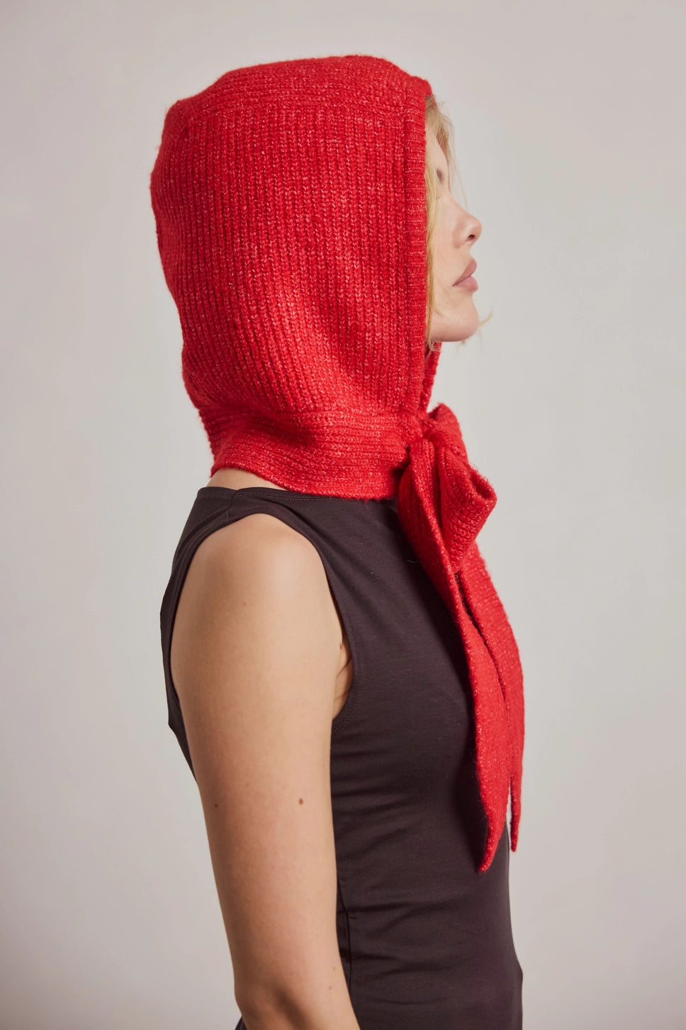 A woman shown in side profile wearing a bright red textured knit bonnet with a long tied tail, over a dark sleeveless top, against a plain light gray background.
