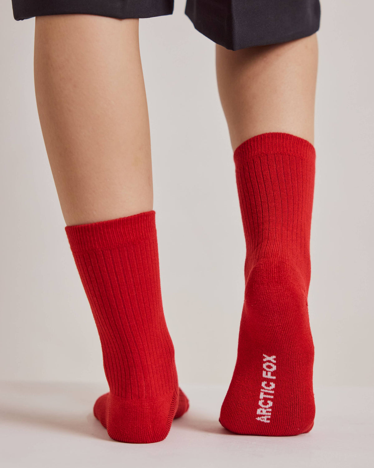 A person wearing bright red crew socks with the brand name 'ARCTIC FOX' printed in white on the sole of one sock, shown from the back against a light gray studio background.