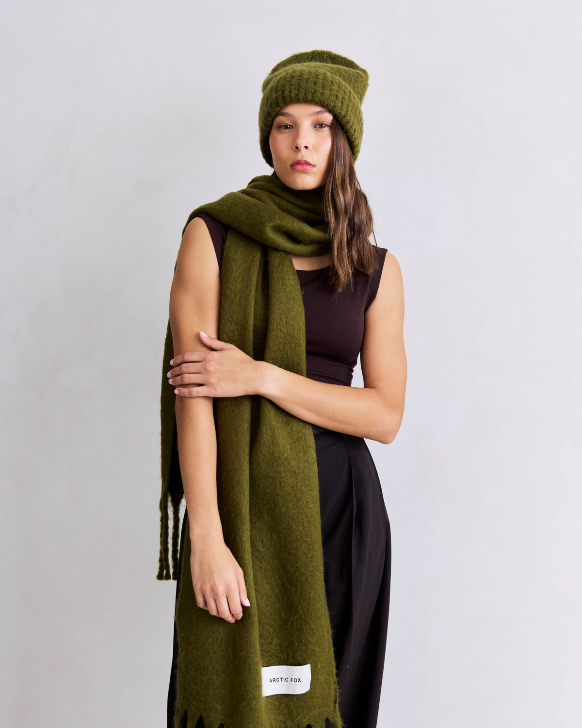 A woman modeling an olive green knit beanie, matching scarf, and a dark brown sleeveless top against a light gray studio background.