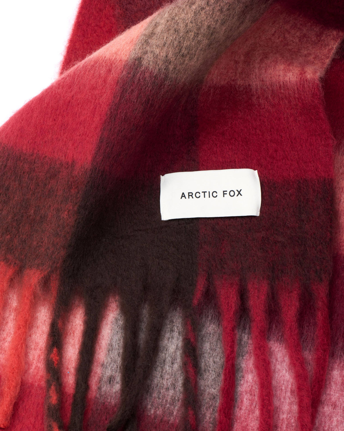 A close-up of a soft plaid scarf in red, dark brown, and black, with fringe at the bottom and a white Arctic Fox brand label sewn onto the fabric.