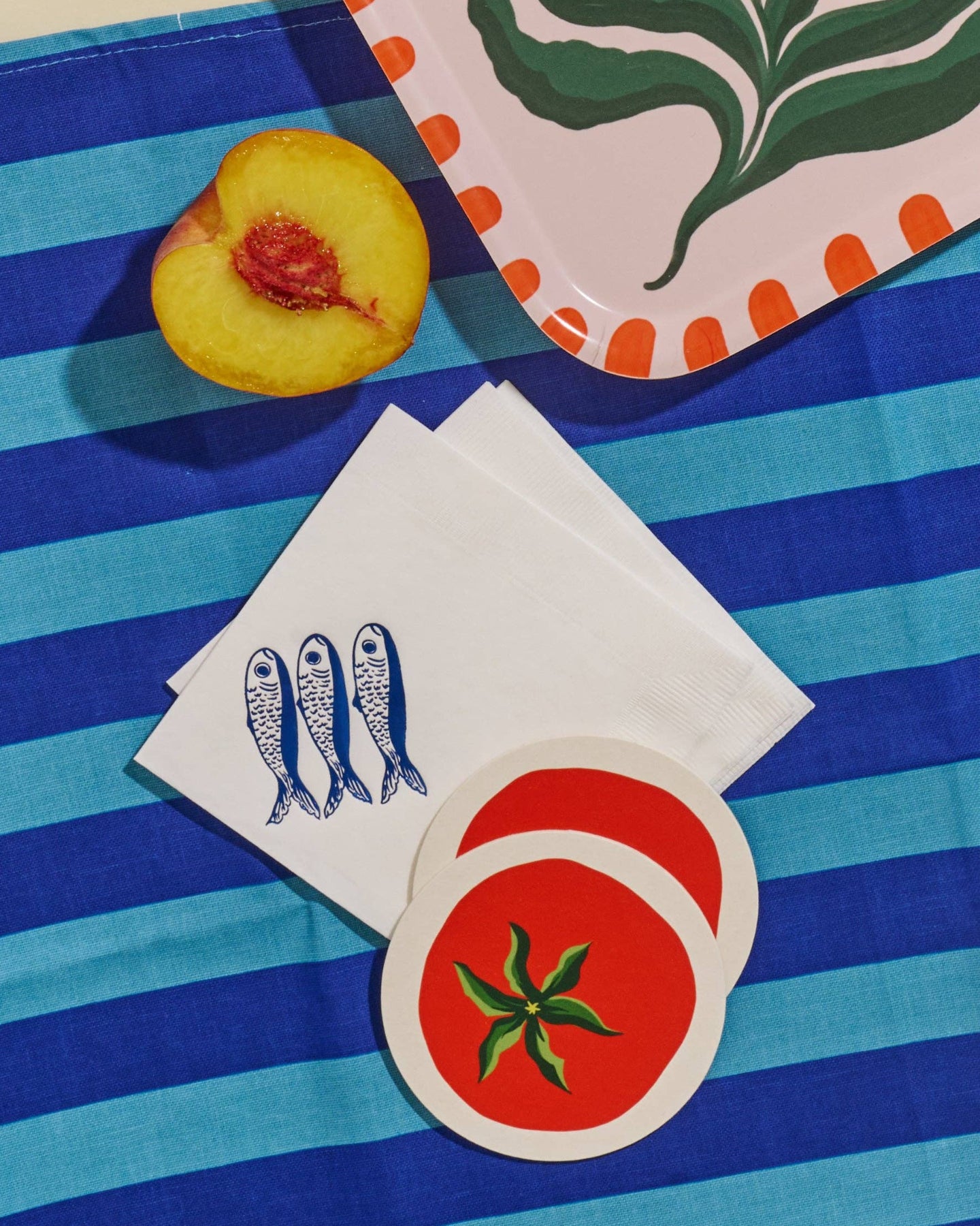 Stack of white sardine napkins, tomato coasters, and peach on a blue and white striped surface.