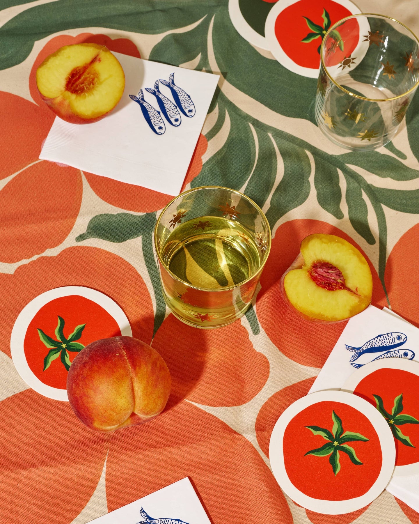 Table setting with peaches, a glass of white wine, and coasters on a patterned tablecloth.