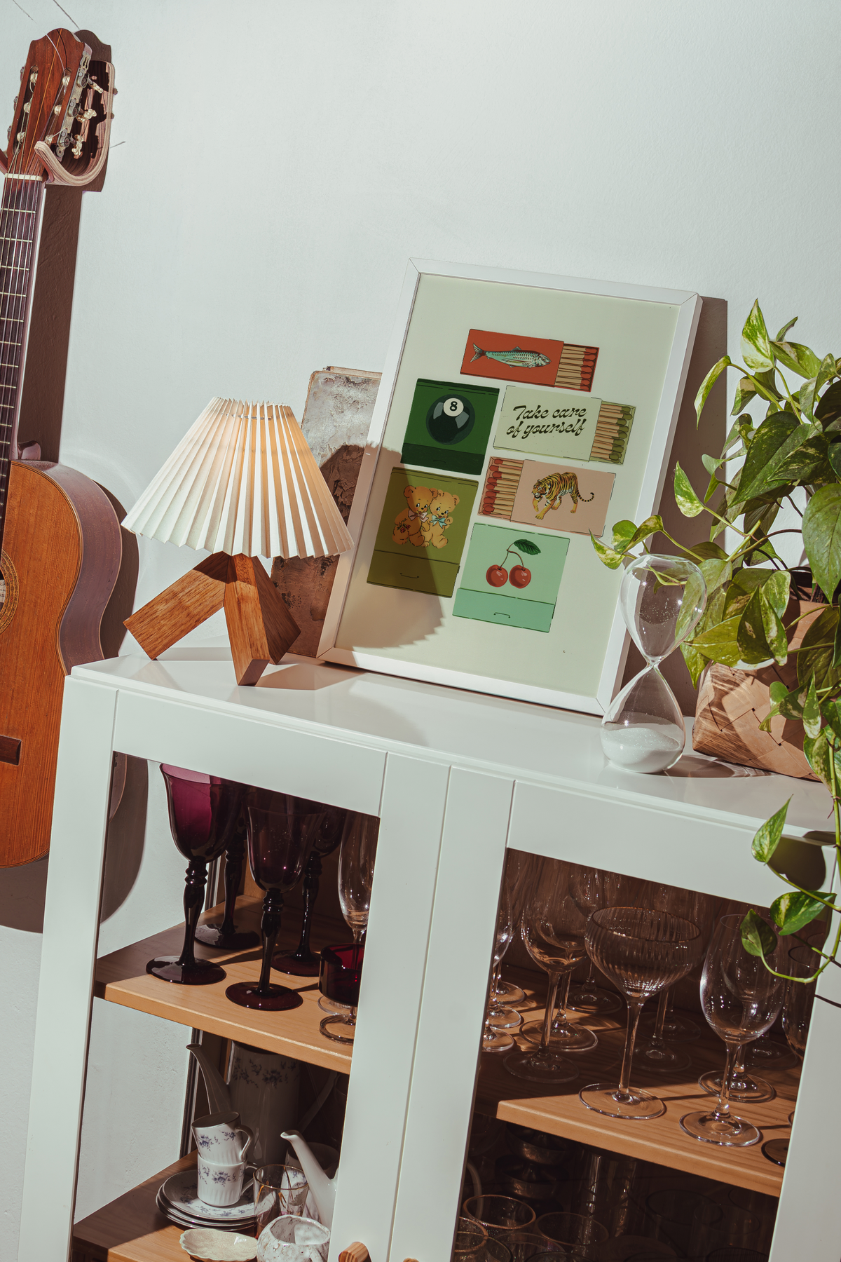 A framed art print with retro-style colorful illustrations and text sits on a white cabinet in a styled room setting, alongside a pleated lamp, wine glasses, a guitar, and a potted plant.
