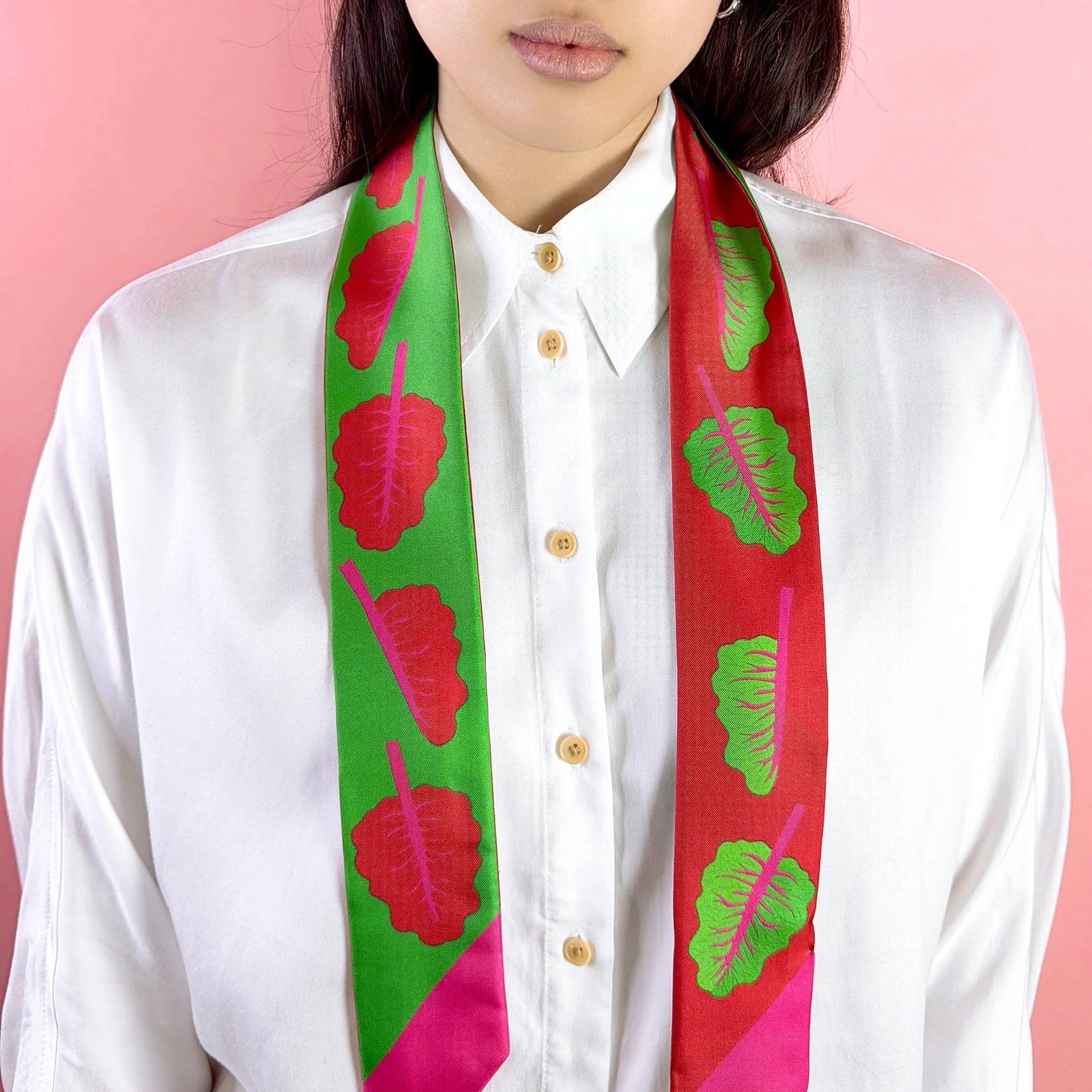Person wearing a white shirt with a colorful scarf featuring red and green patterns on a pink background, twilly-ribbon-silk-scarf-red-chard-green-centinelle-02