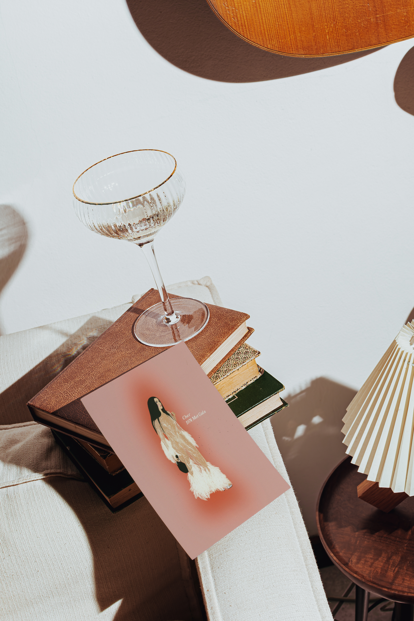 A styled interior scene with a pink ART PRINT OF CHER AT THE MET GALA featuring a white and black bird illustration, resting on a stack of books beside a clear stemmed glass with a gold rim. A pleated cream lamp and warm wood furniture are visible in the softly lit room.