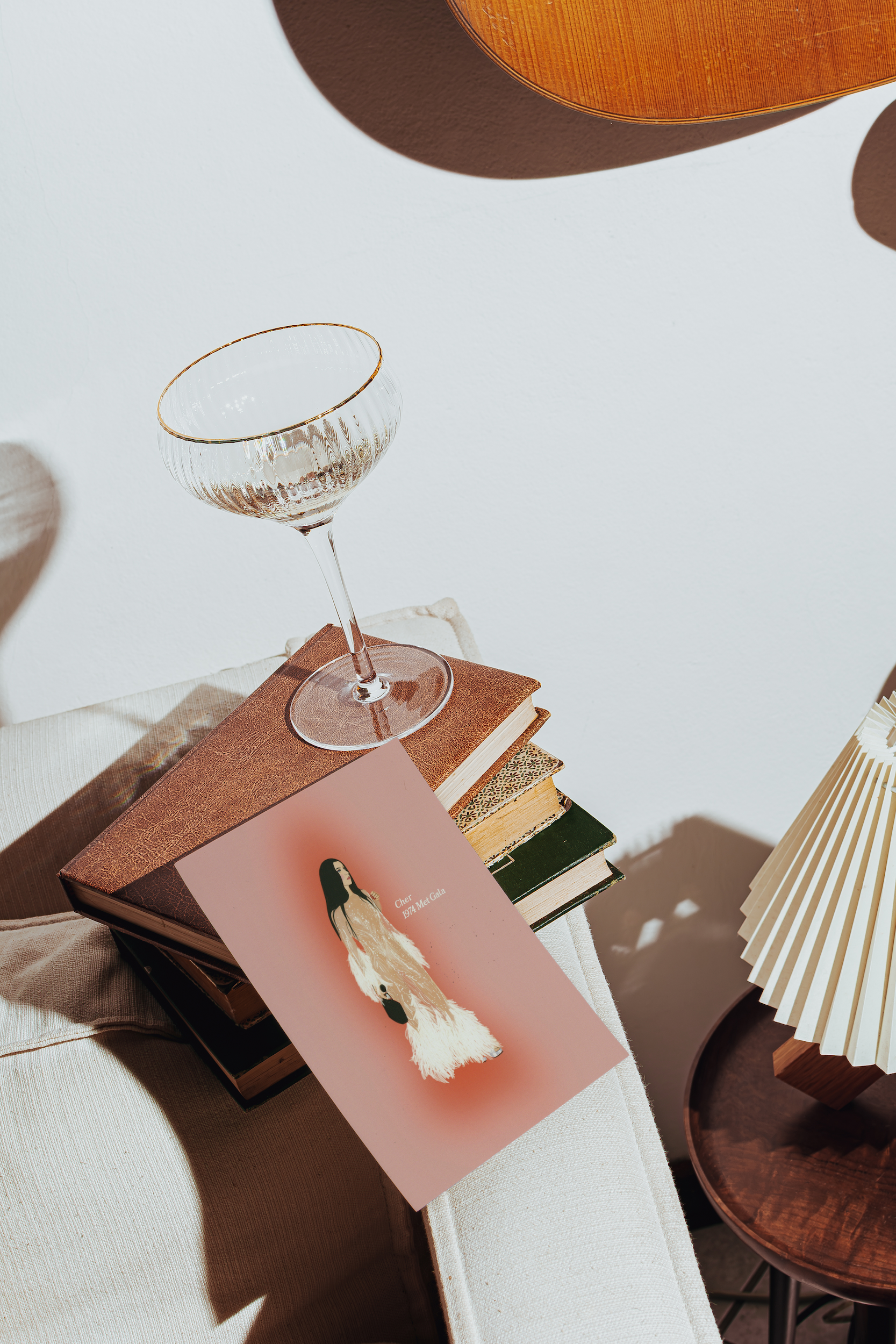 A styled interior scene with a pink ART PRINT OF CHER AT THE MET GALA featuring a white and black bird illustration, resting on a stack of books beside a clear stemmed glass with a gold rim. A pleated cream lamp and warm wood furniture are visible in the softly lit room.