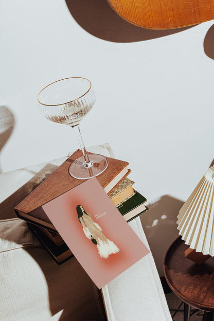 A styled interior scene with a pink ART PRINT OF CHER AT THE MET GALA featuring a white and black bird illustration, resting on a stack of books beside a clear stemmed glass with a gold rim. A pleated cream lamp and warm wood furniture are visible in the softly lit room.