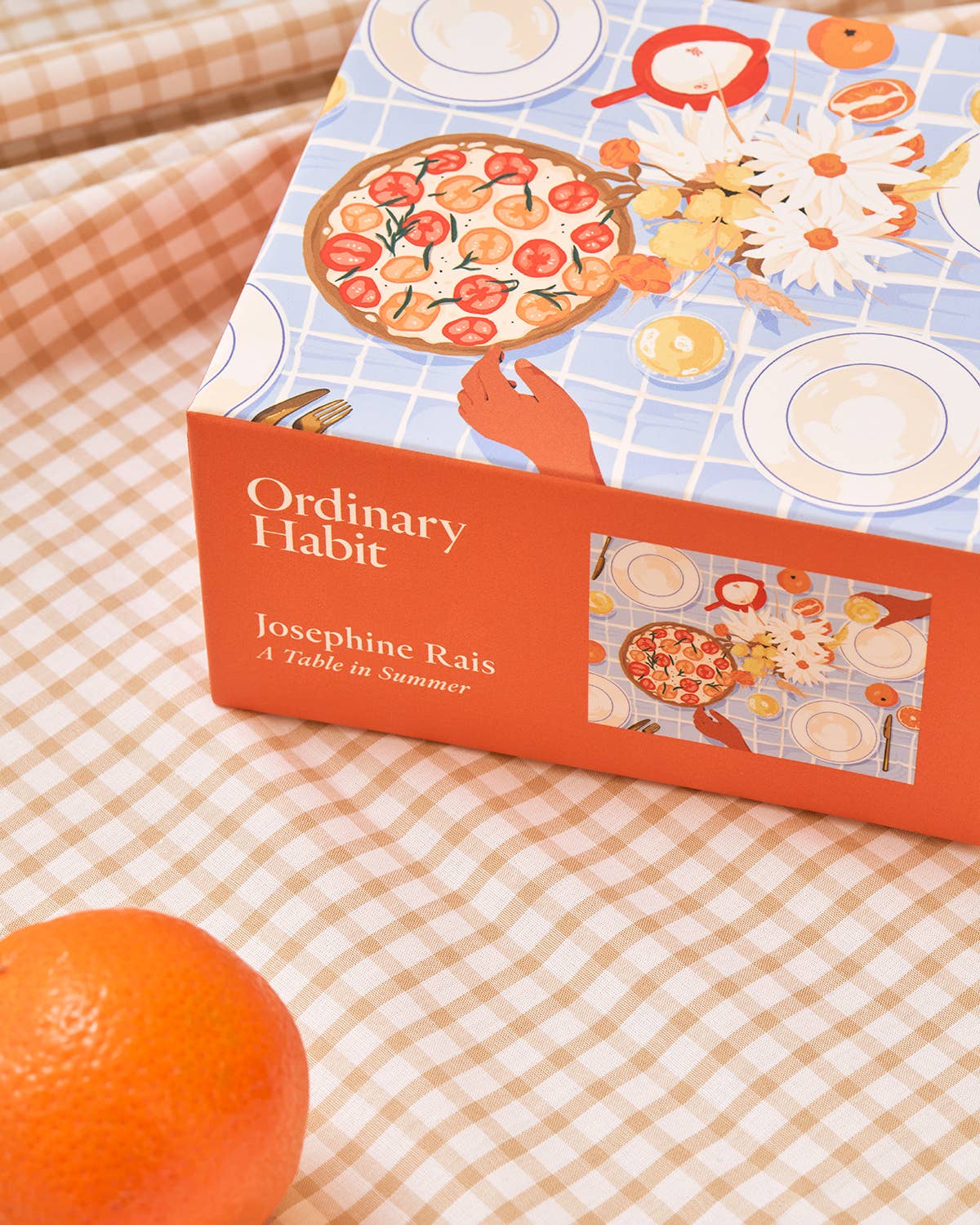 Book titled 'Ordinary Habit' by Josephine Rais on a checkered tablecloth with an orange.