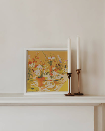 Framed artwork with flowers on a shelf next to two white candles in bronze candle holders.