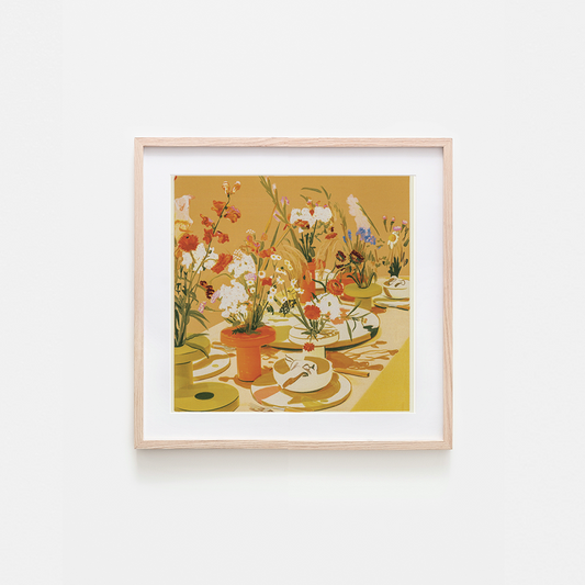 A framed art print featuring a colorful still life illustration of a table filled with flowers, plates, and cups, set against a warm ochre background. The print is housed in a light natural wood frame with a white mat, mounted on a clean white wall.