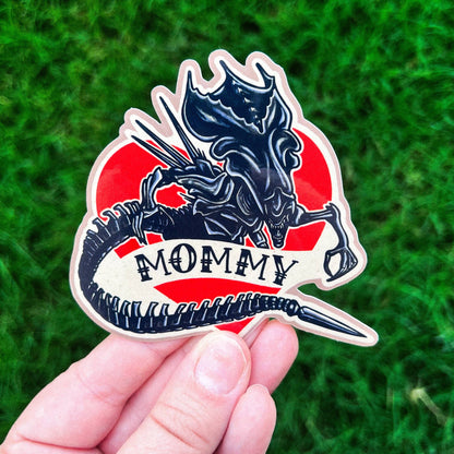Sticker with a dragon design and 'Mommy' text held against a grass background