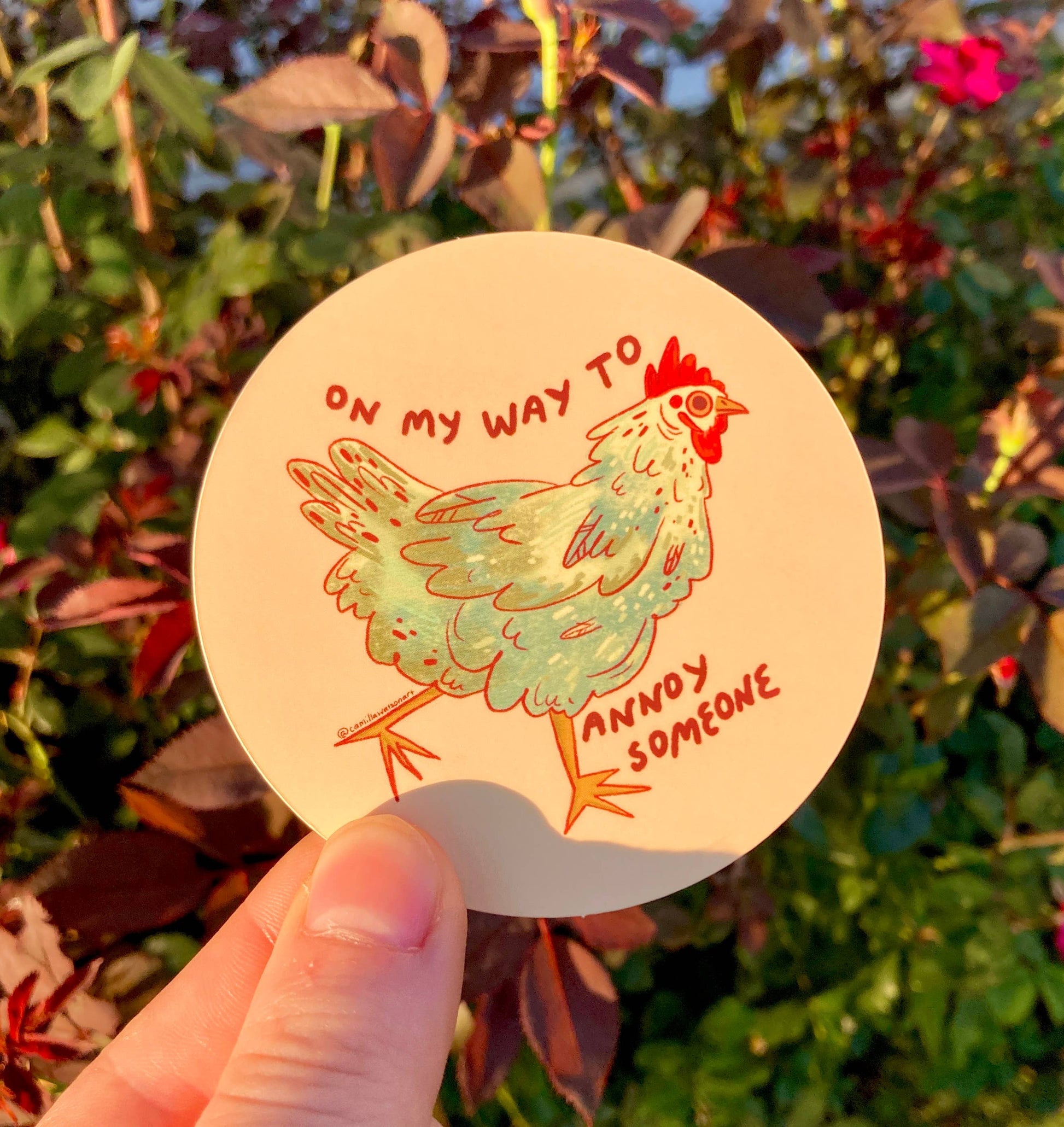 A hand holds a round sticker featuring a cartoon chicken with the text 'ON MY WAY TO ANNOY SOMEONE' against a blurred garden background.