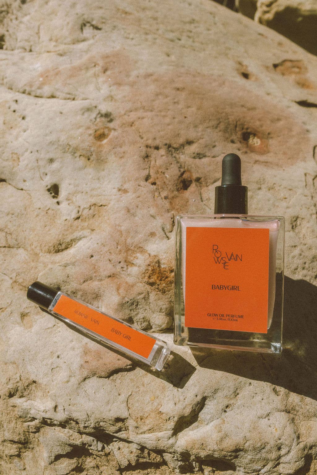 A clear glass perfume bottle with a black dropper cap and an orange label reading 'RIVIERA' sits on a sunlit rock next to a matching orange-and-clear spray vial.