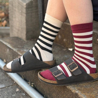 A close-up of a person sitting on stone steps wearing two pairs of striped crew socks and dark brown double-strap slide sandals. One sock has black and cream stripes, and the other has burgundy and cream stripes.