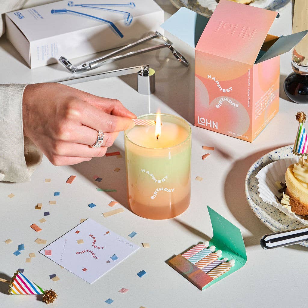 Person lighting a birthday candle on a colorful candle with confetti and a cake in the background.