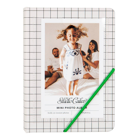 A mini photo album with a white and black grid pattern cover, featuring a sample photo of a young girl and the text 'Stille Källo Mini Photo Album'. A green pencil is placed diagonally across the front of the album against a plain white background.