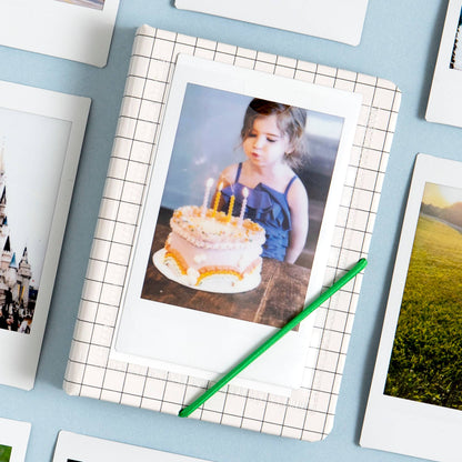 A square photo print with a white border showing a young girl blowing out birthday candles on a cake, placed on a white-and-gray grid-patterned mat. A green pencil rests across the print, and several other framed or bordered photos are partially visible around it on a light blue surface.